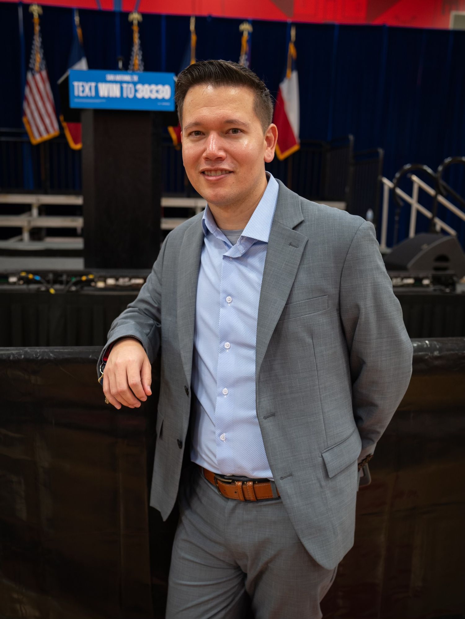 A person in a gray suit and light blue shirt stands in front of a dark backdrop at an event.