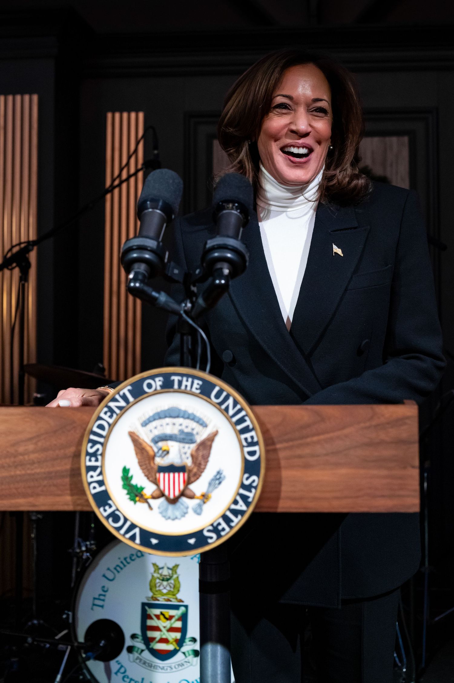 A person in a black coat and white shirt stands behind a podium with the United States Vice Presidential seal.