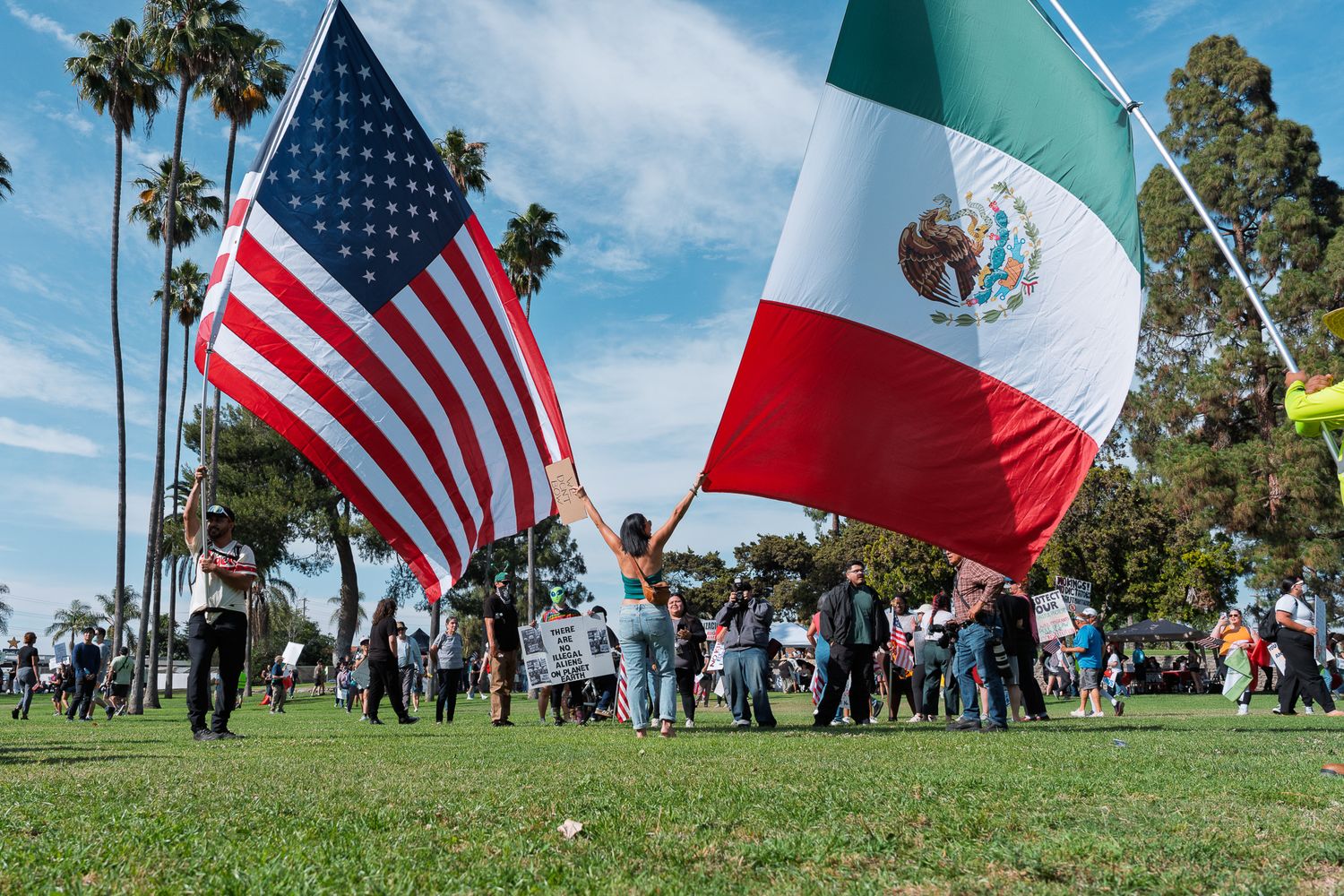 People gathered outdoors holding American and Mexican flags against blue sky with palm trees.