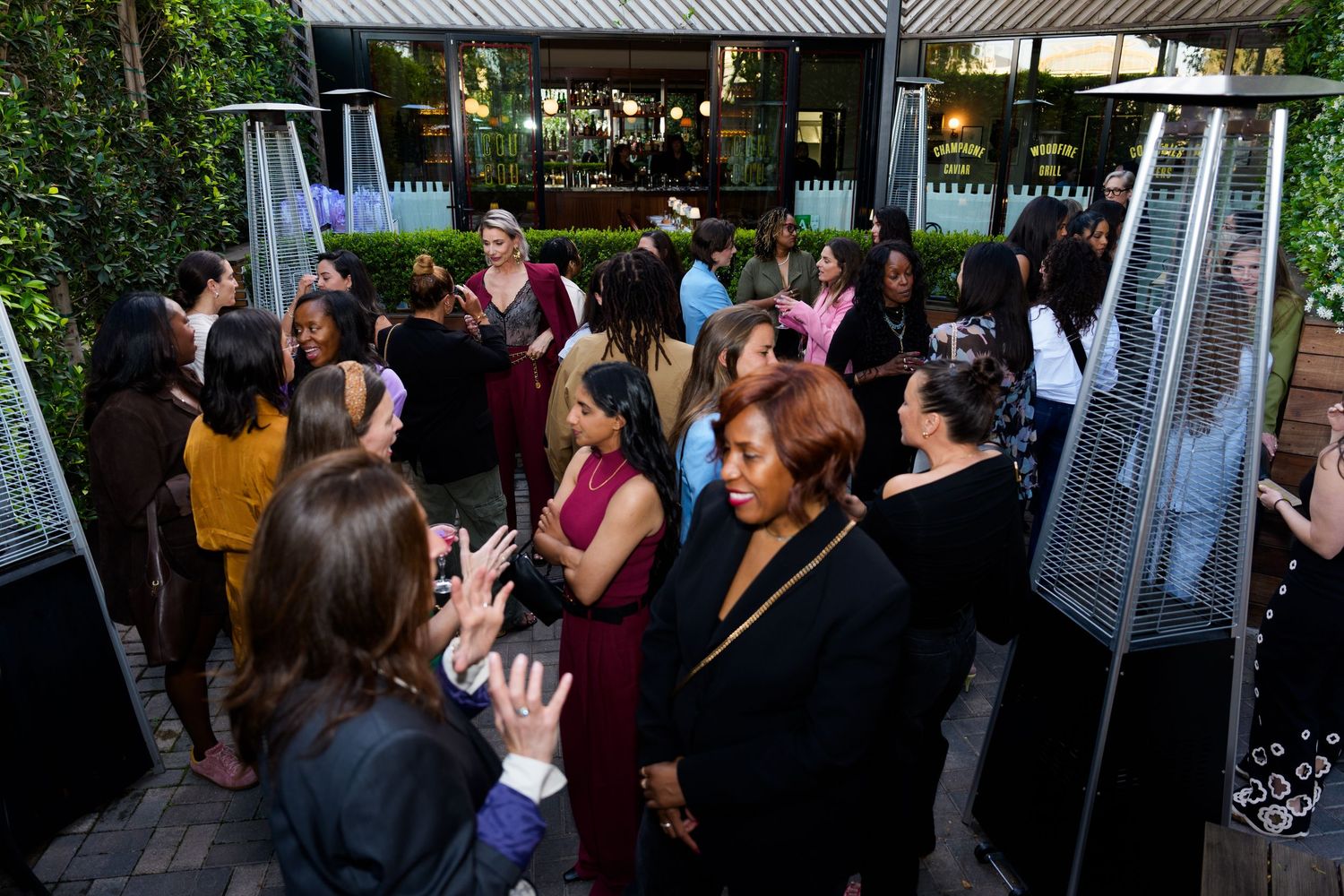 Diverse group of elegantly dressed women networking at an upscale outdoor evening event with lush greenery.
