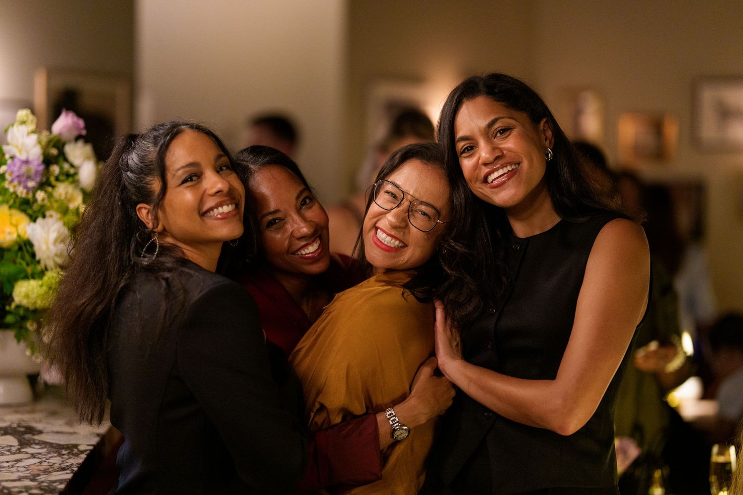Four women smiling and hugging together at an indoor social gathering with flowers in the background.