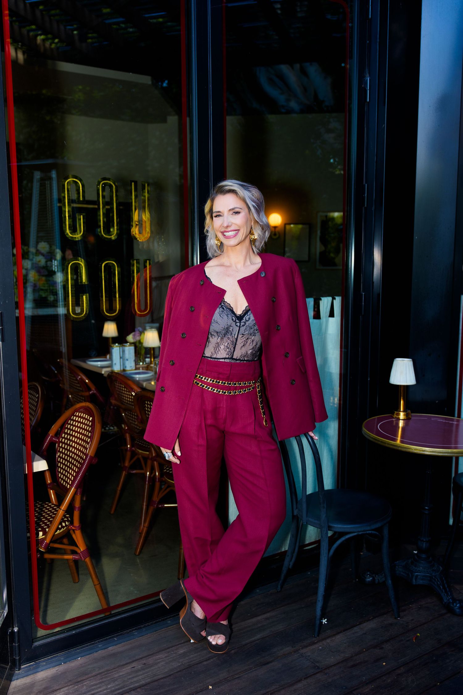 Woman in burgundy suit poses outside Coucou restaurant, wearing lace top and chain belt, smiling confidently.