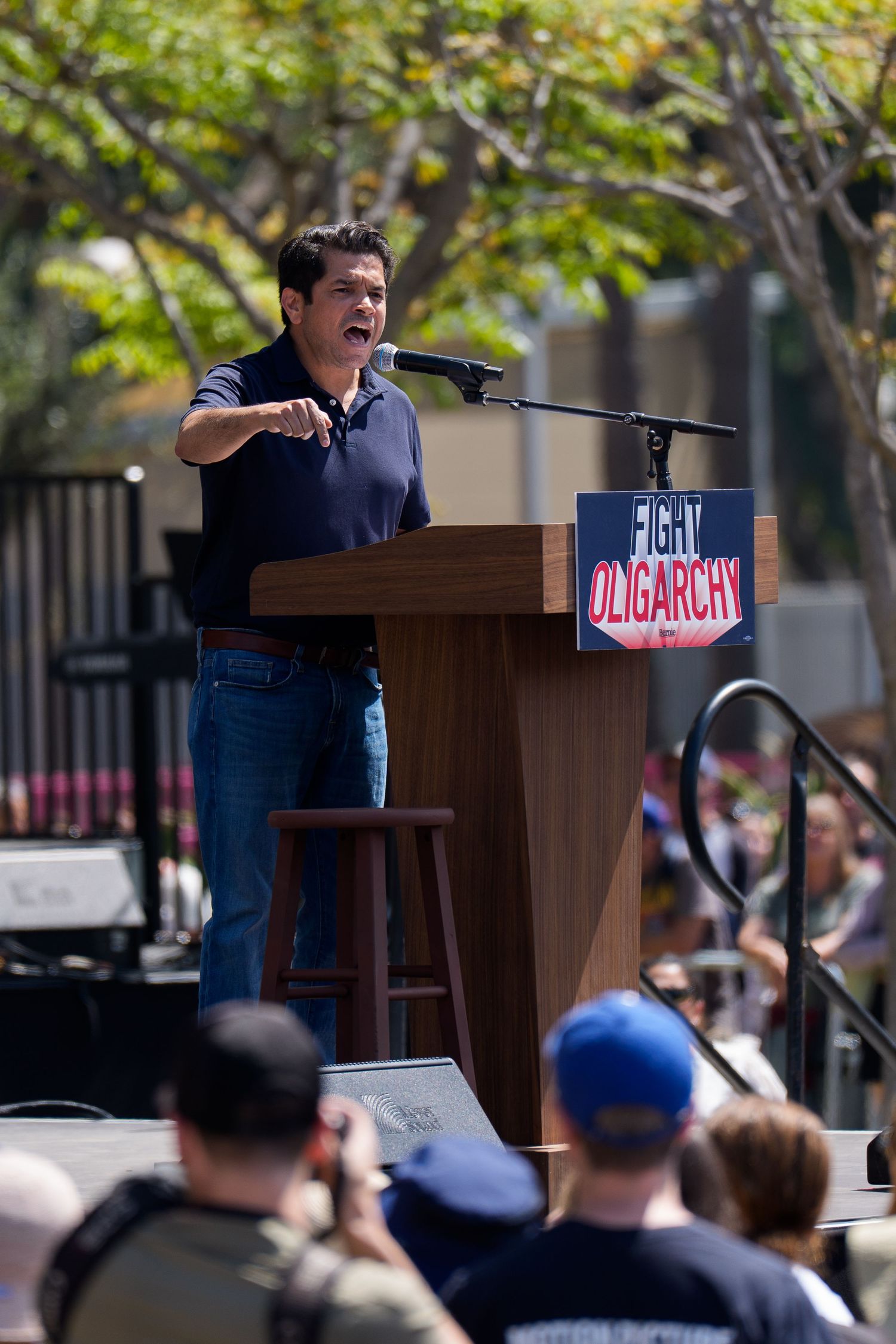 Speaker gesturing while delivering remarks at outdoor podium with campaign signage.