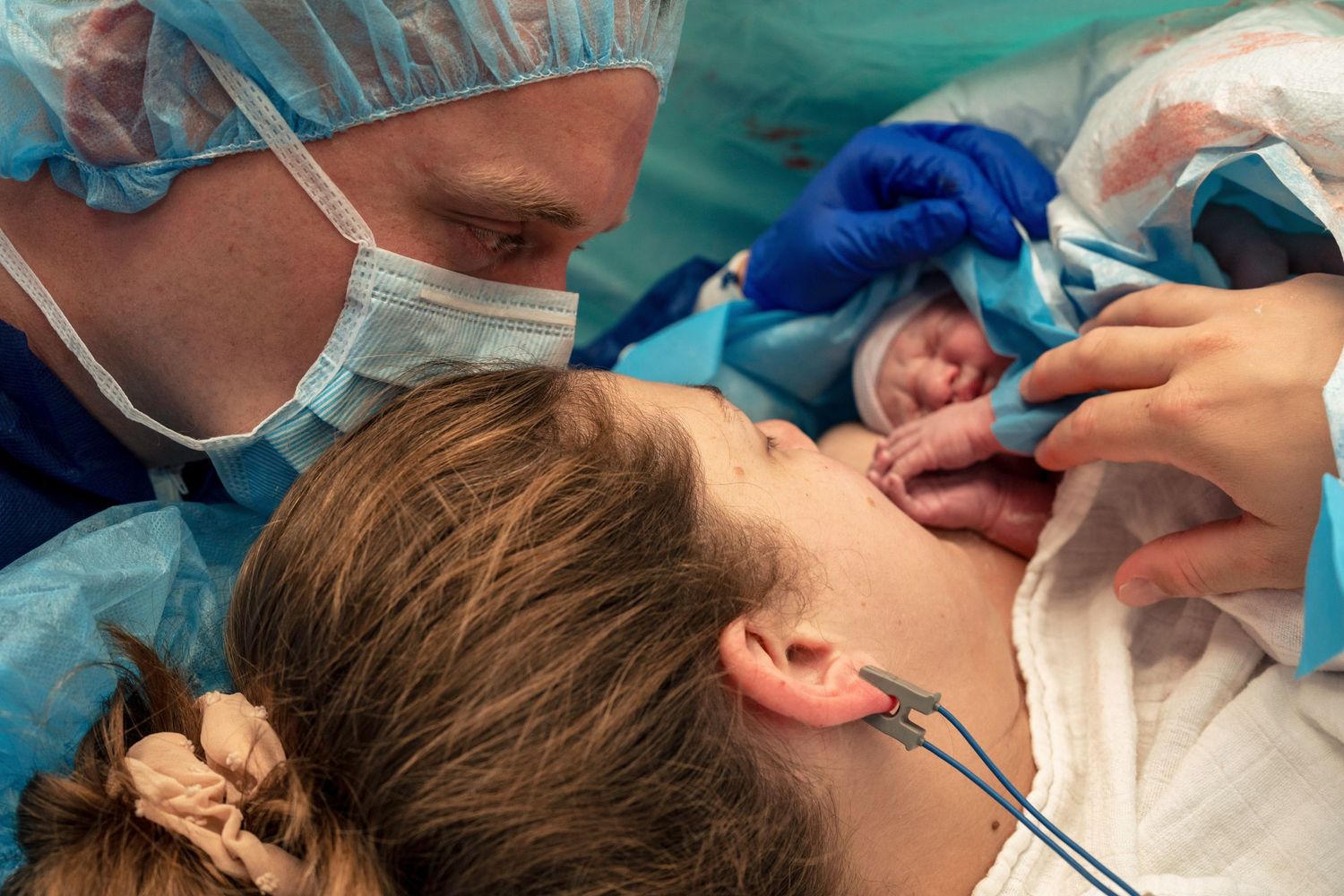 Doctor in mask cuts umbilical cord as mother bonds with newborn baby after C-section delivery. Birth photography by Constanze Ariane