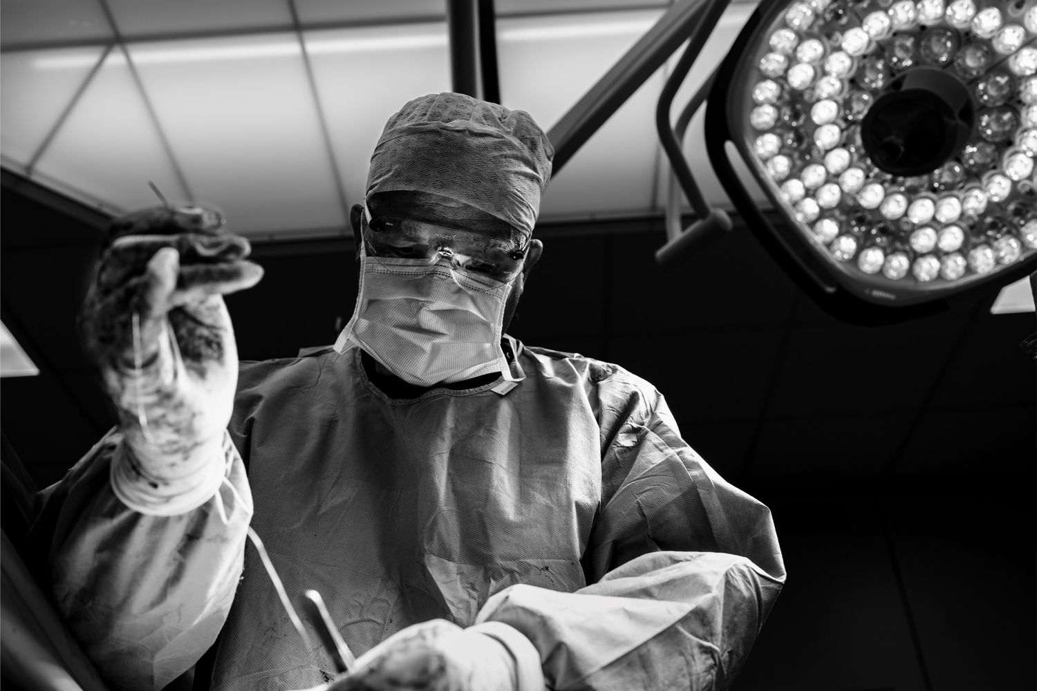 Black and white photo of a surgeon in scrubs and mask under bright operating room lights suturing. Medical documentary by Constanze Ariane