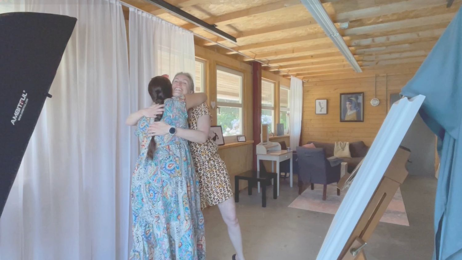 Two women in floral dresses embracing in a bright studio space with wooden ceiling and elegant decor.