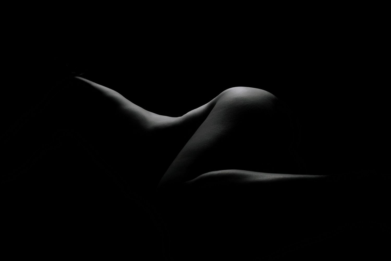 Black and white fine art nude photography showcasing elegant body curves against a dark background.