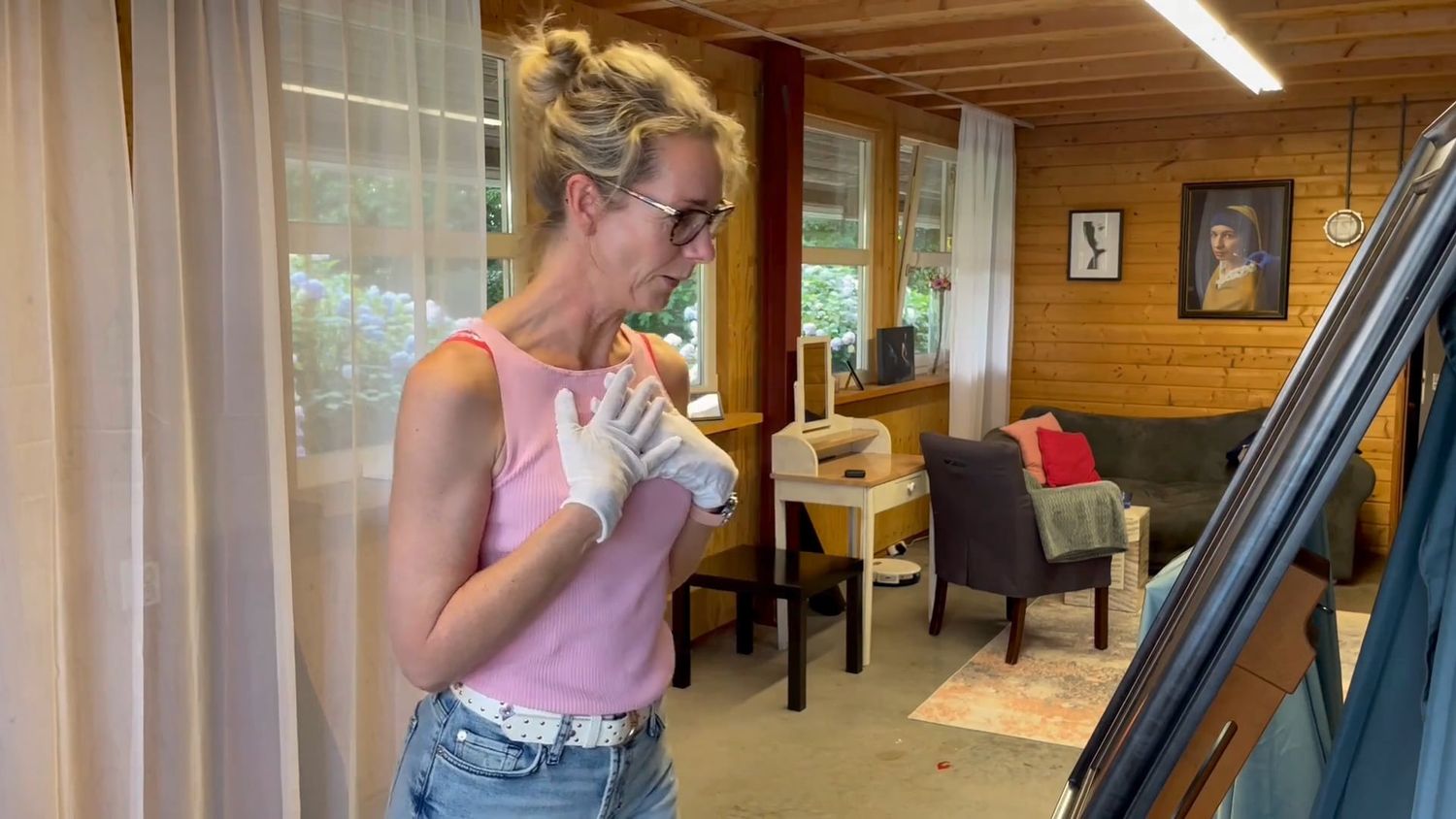 Woman in pink tank top and jeans wearing white gloves working on a project inside a cozy wooden cabin studio.
