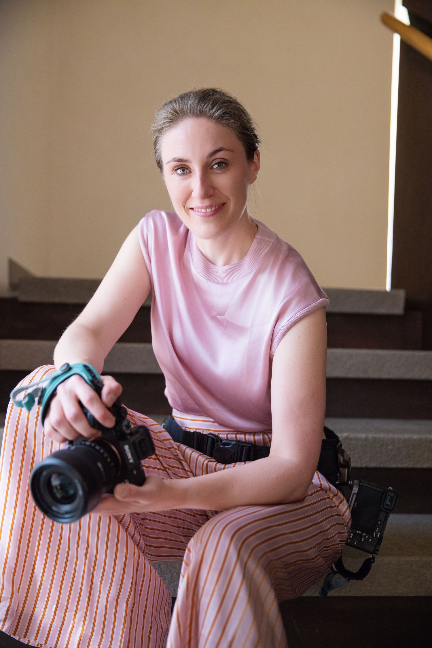 Smiling portrait of Constanze Ariane seated on stairs holding a professional camera in pink outfit.