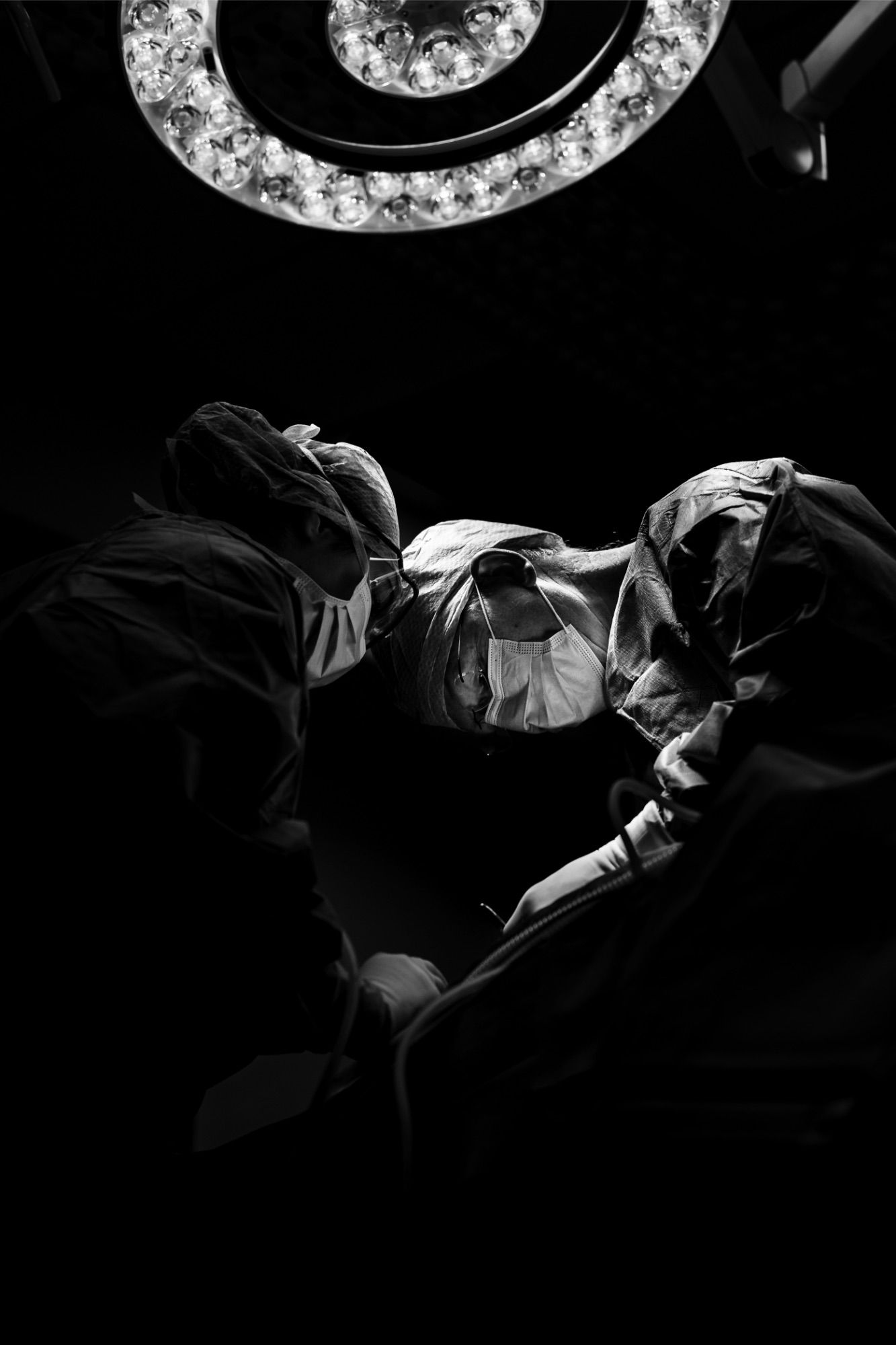 Dramatic black and white silhouette of surgeons operating under a circular surgical light in a dark operating room.
