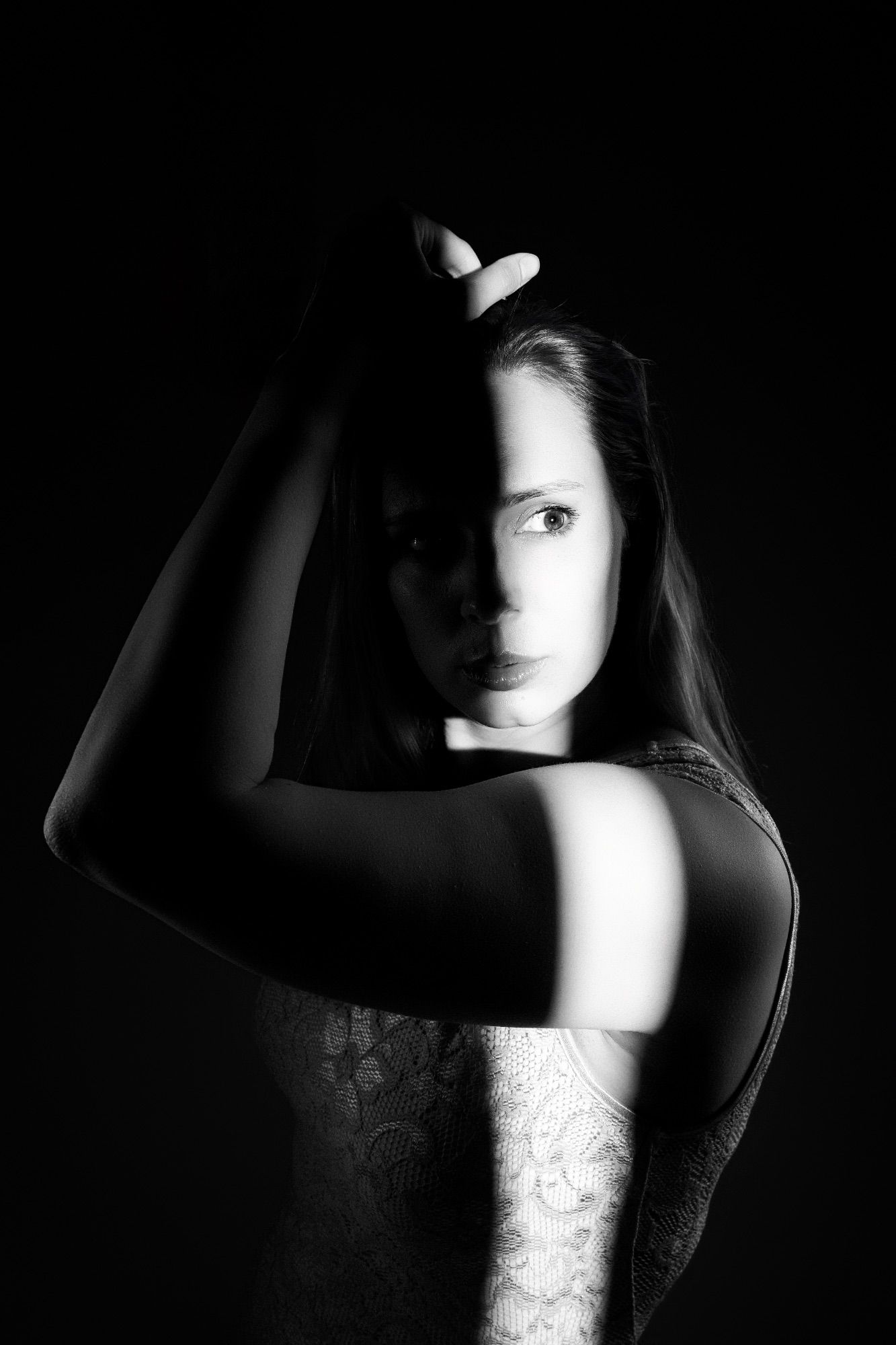 Black and white portrait of a young woman with hand raised above head, dramatic shadow lighting on dark background. Editorial fine art portr