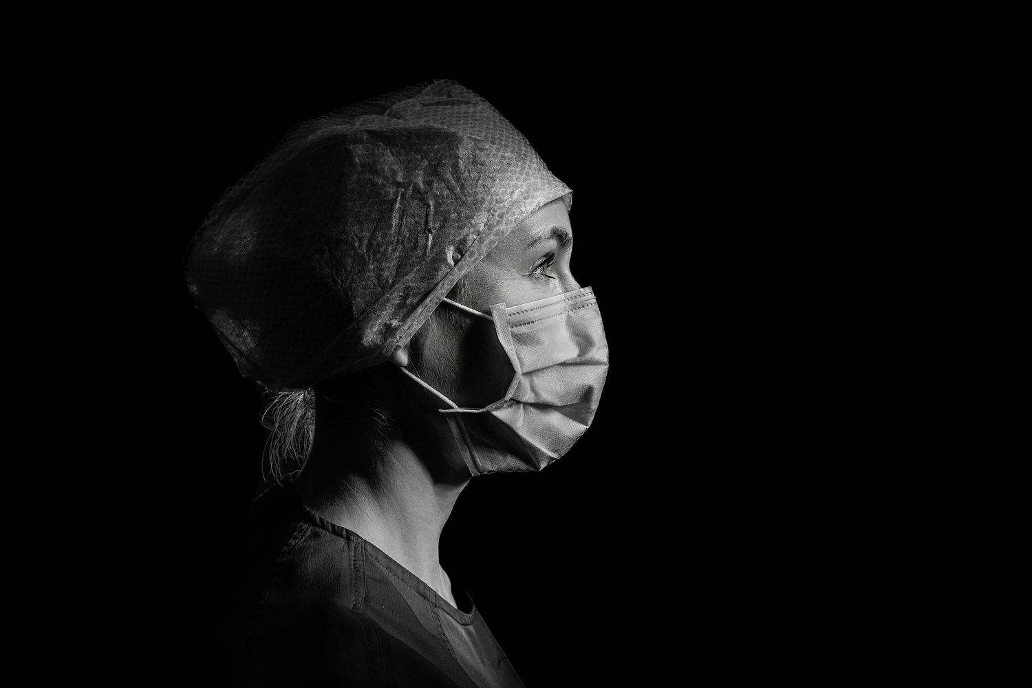 Black and white profile portrait of a female healthcare professional wearing surgical mask and cap against dark background.