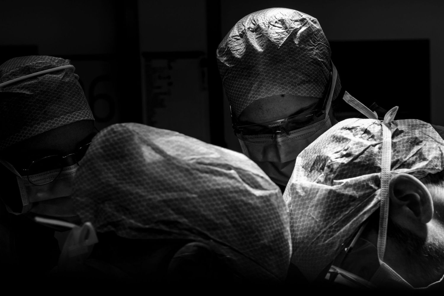 Two masked surgeons working closely together under bright operating lights during a medical procedure.