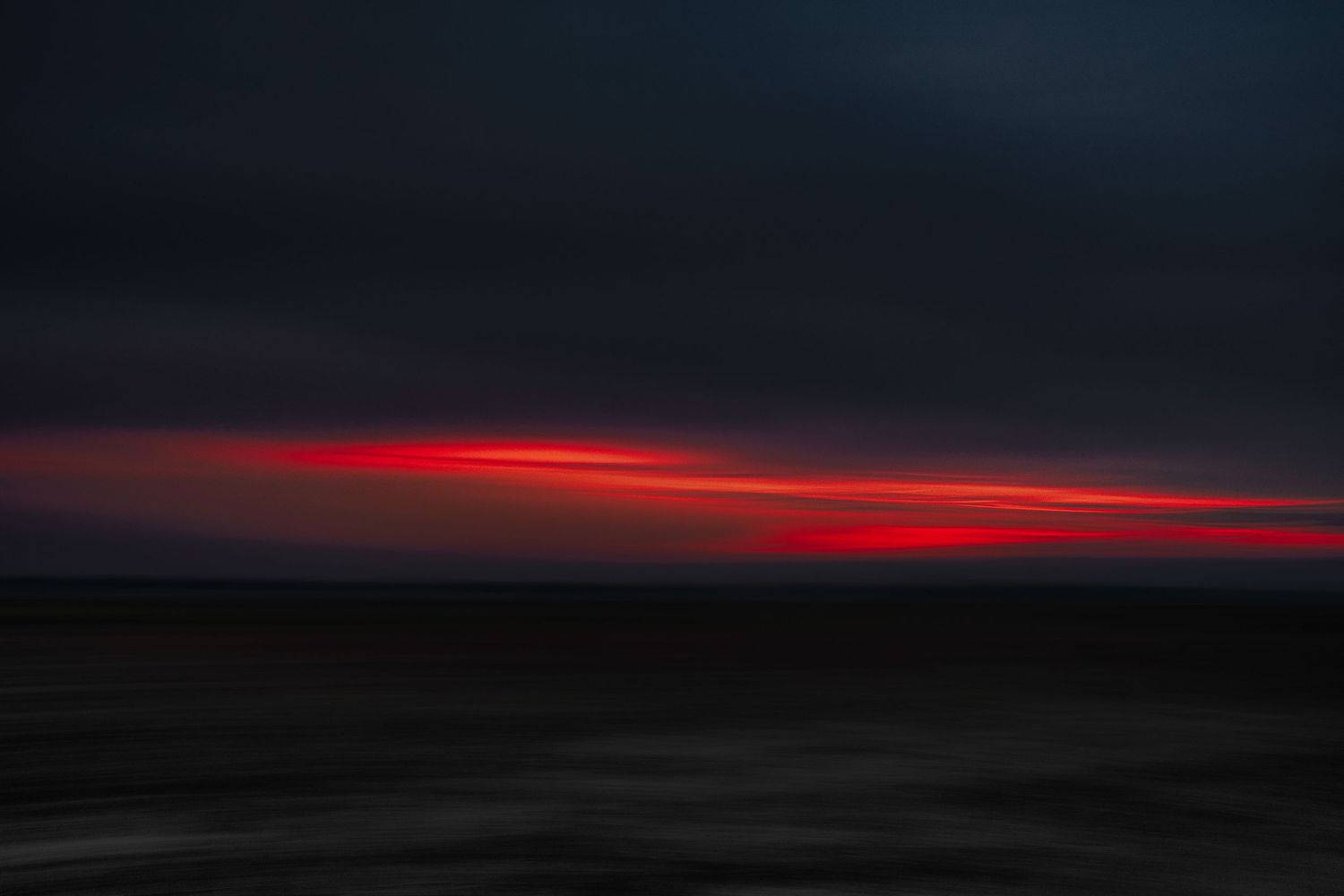 Dark seascape at sunset with dramatic red and orange horizon glowing against stormy dark clouds over calm ocean waters. ICM photography.