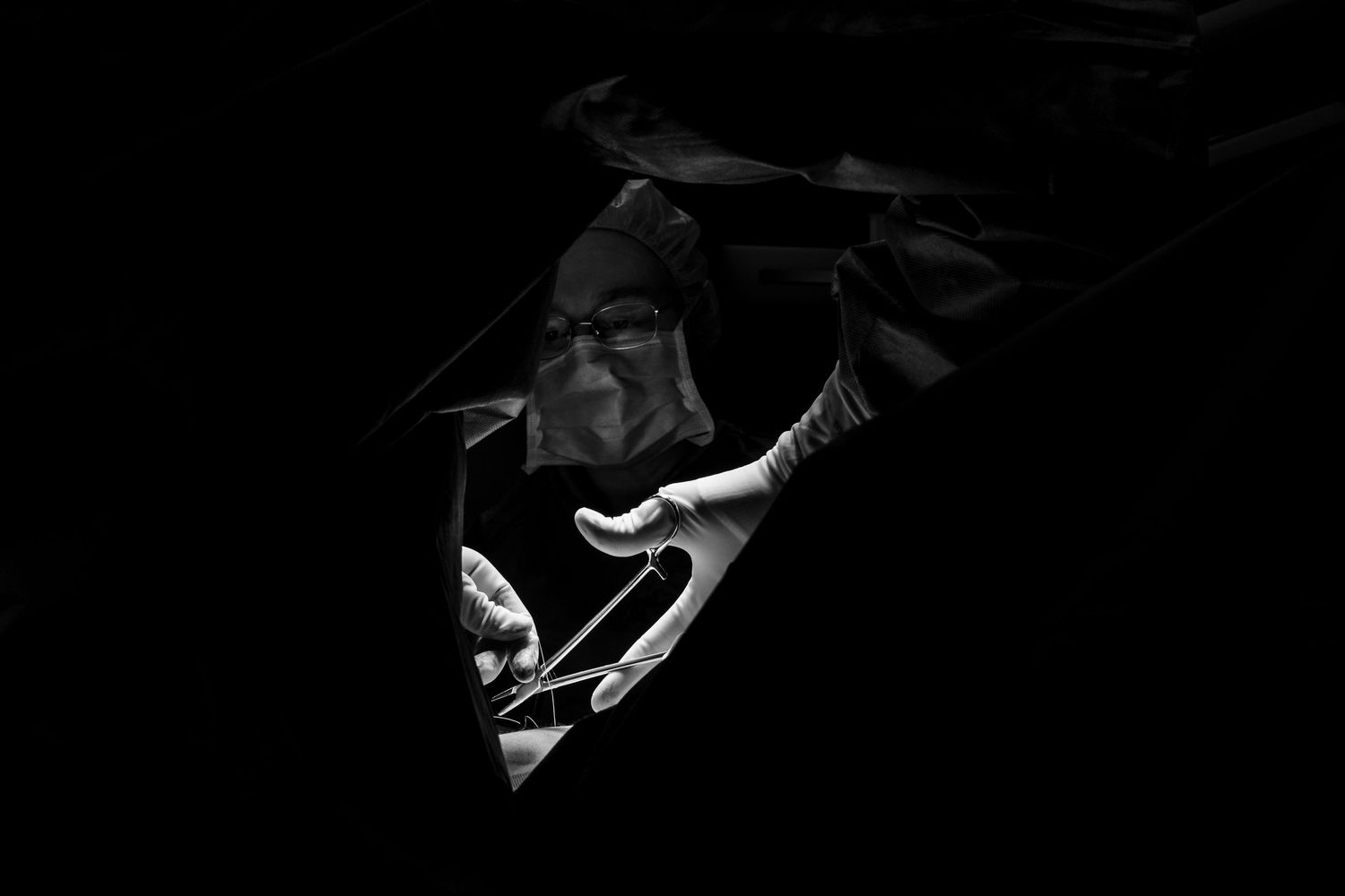 Dramatic black and white portrait of a masked surgeon illuminated by operating light holding surgical scissors.