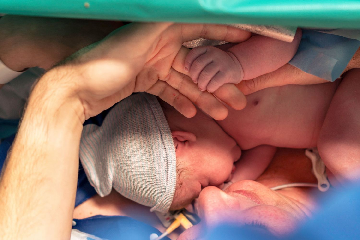 A newborn baby in a hospital cap is held by parents hands during skin-to-skin contact after cesarean birth. Birth photography by Constanze A