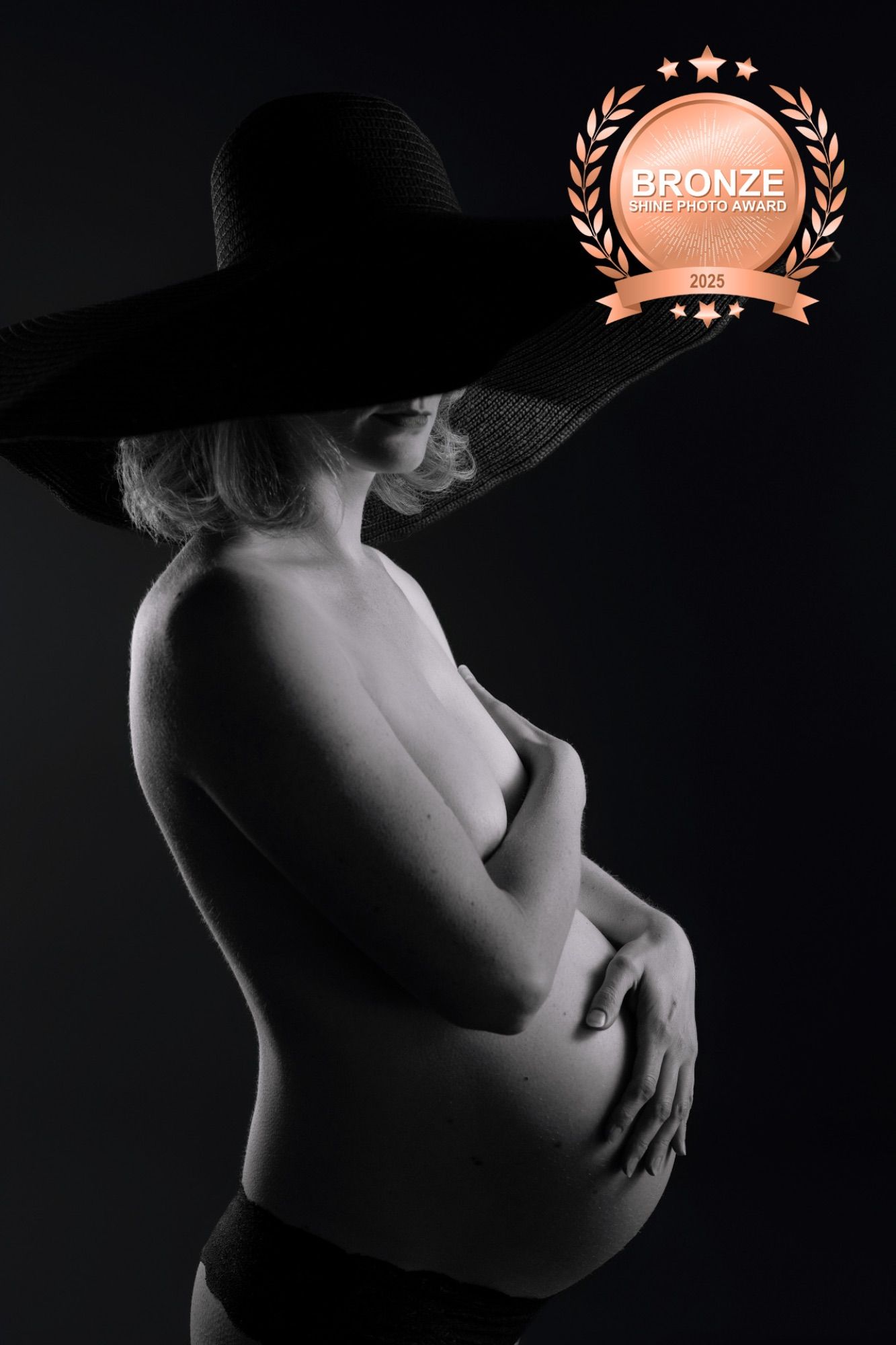 Black and white side profile of a pregnant woman in a wide-brim hat, cradling her baby bump, award-winning maternity photo.
