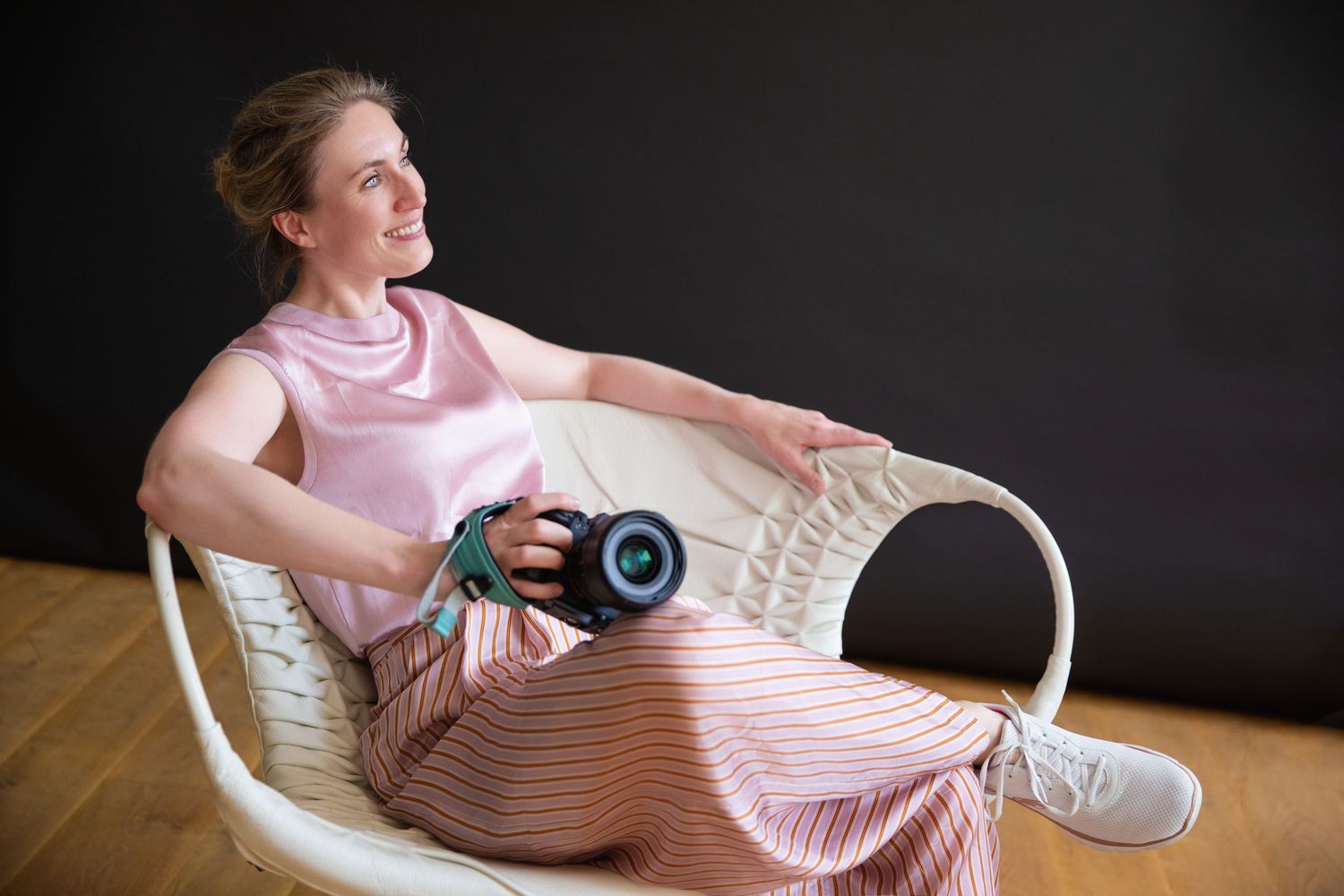 Smiling woman in pink outfit relaxing in wicker chair holding a professional camera against dark background.