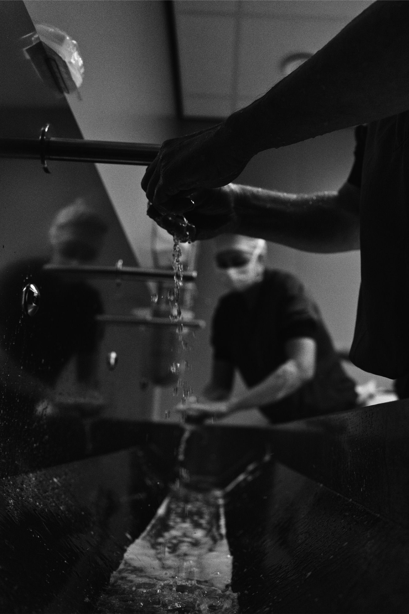 Black and white photo of a masked chef washing hands at a sink, water cascading down, with reflection visible in mirror.