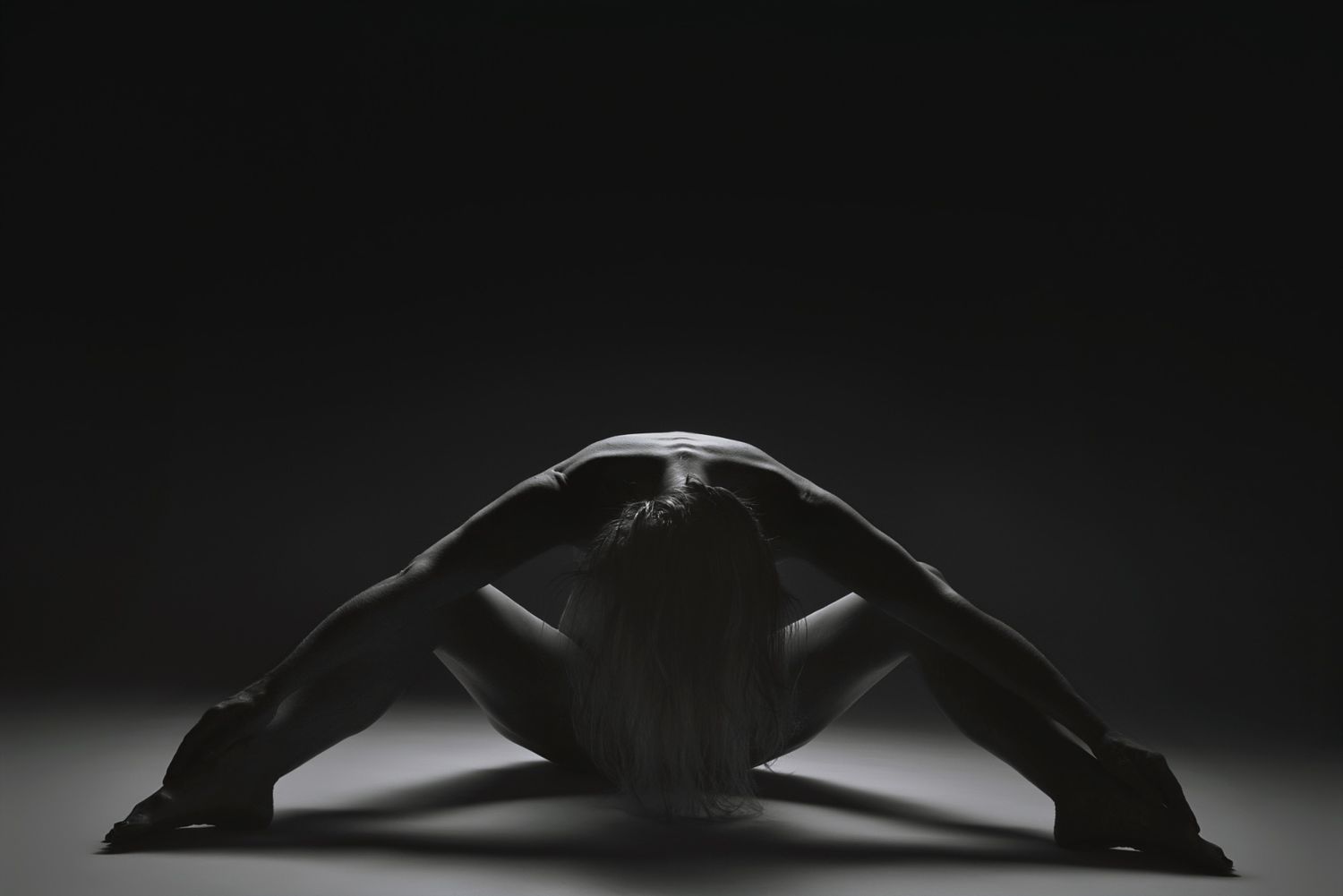 Black and white fine art photo of a flexible dancer in a deep bend pose on a studio floor. Editorial fine art portrait by Constanze Ariane