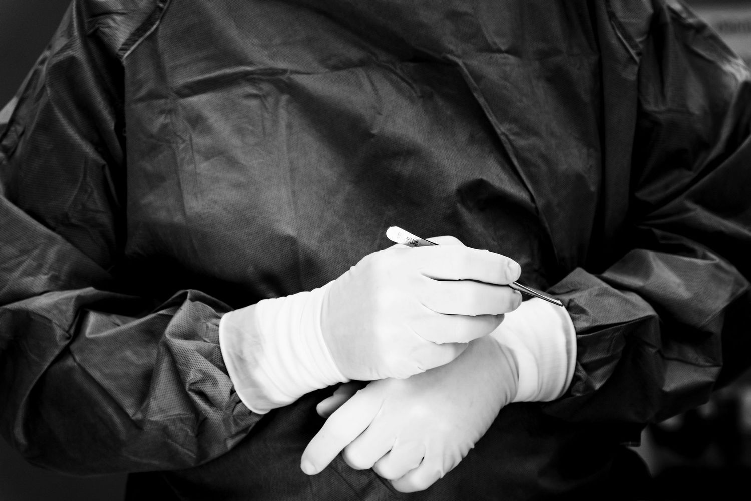 Surgeon in black gown wearing white latex gloves clasped together in operating room. Medical documentary by Constanze Ariane