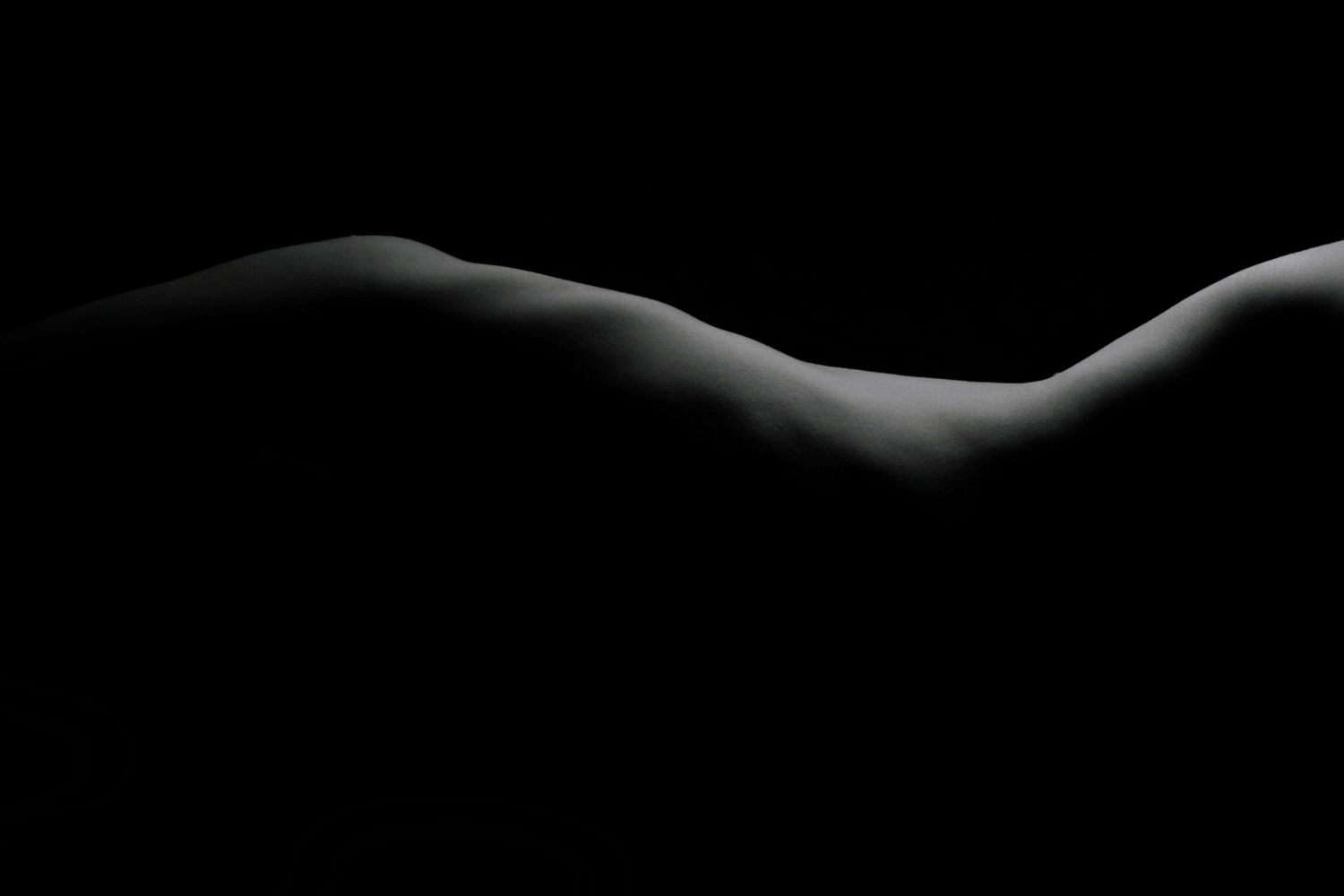 Black and white fine art nude bodyscape photograph highlighting curves and shadows against a dark background.