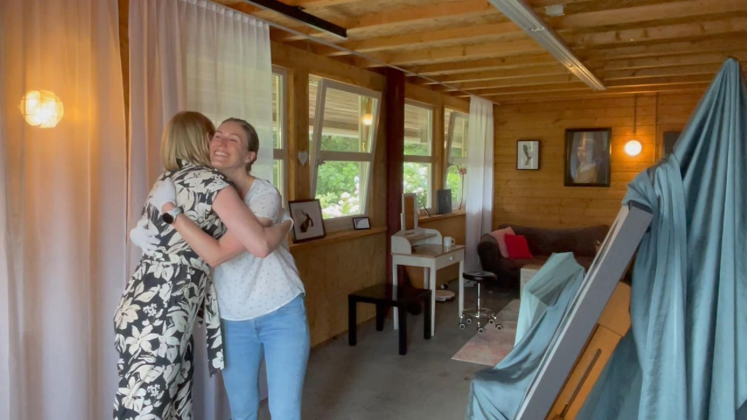 Two people embrace in a cozy wooden cabin interior with large windows, curtains, and rustic furnishings.