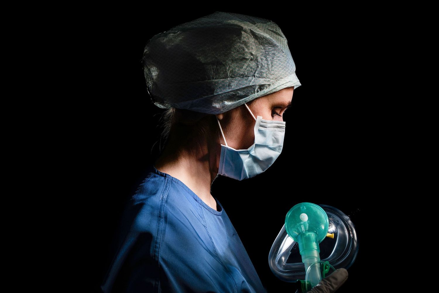 A medical professional in blue scrubs, surgical cap, and face mask holds an oxygen mask against a dark background.
