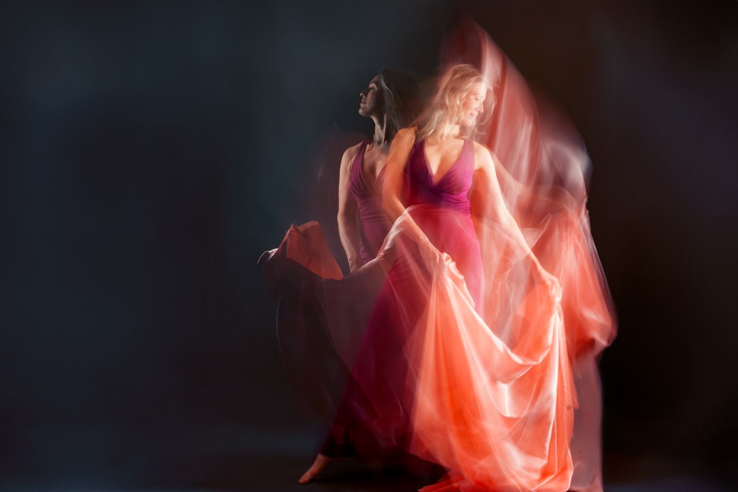 A blurred motion photo of a woman in a flowing red and pink gown dancing gracefully against a dark background.