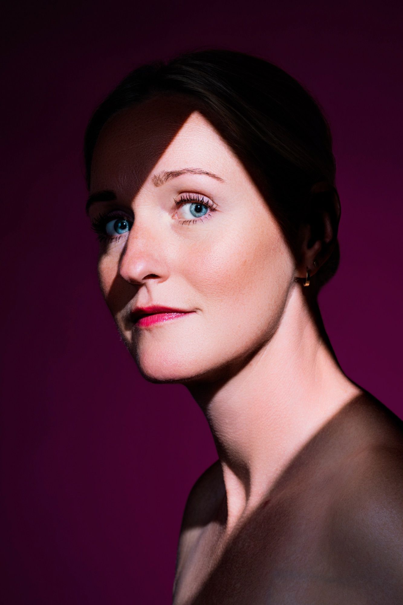 Woman with blue eyes and red lips poses against a purple background in dramatic split lighting portrait by Constanze Ariane fine art
