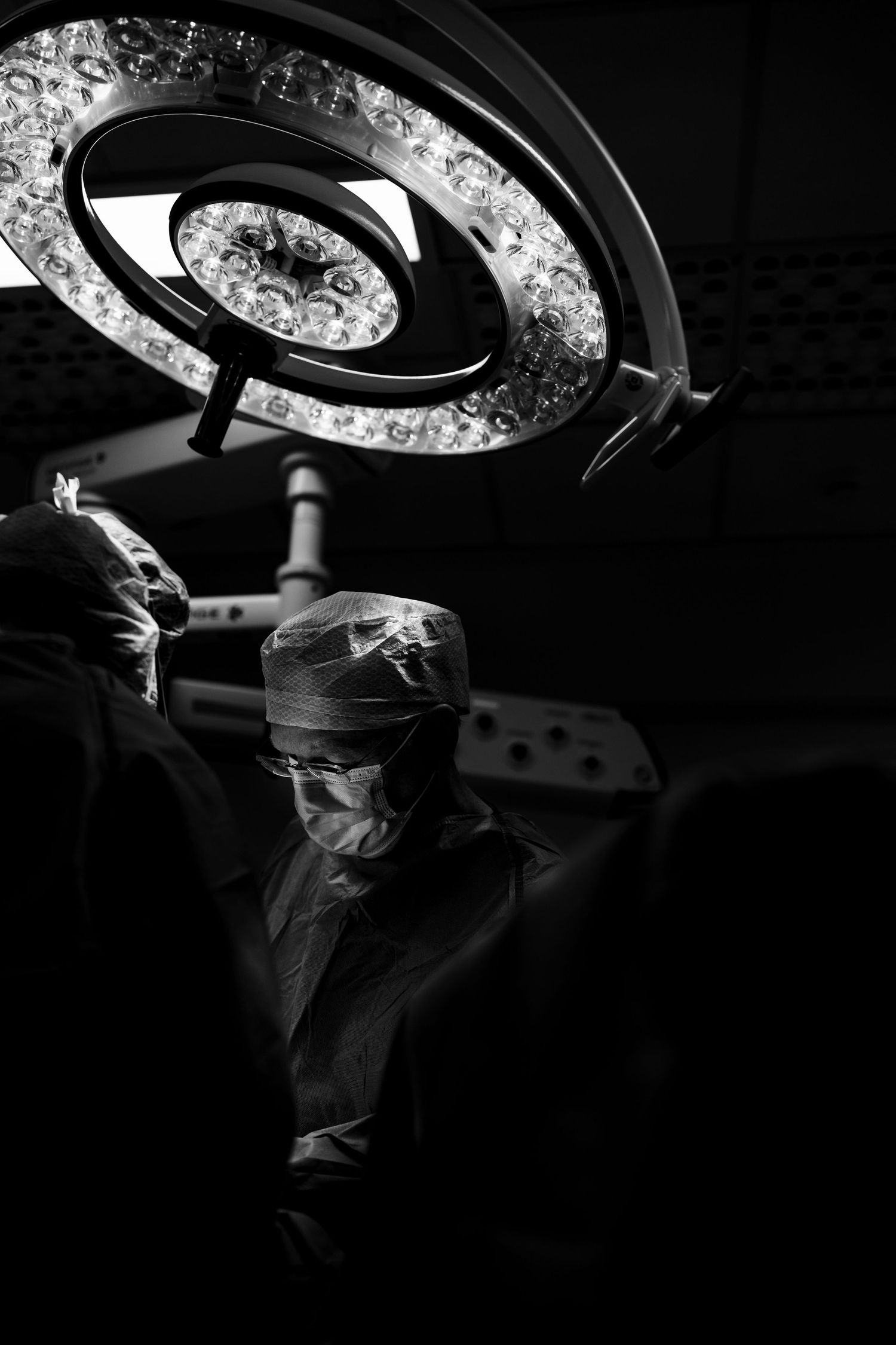 Two surgeons in masks beneath bright circular surgical light in dark OR.