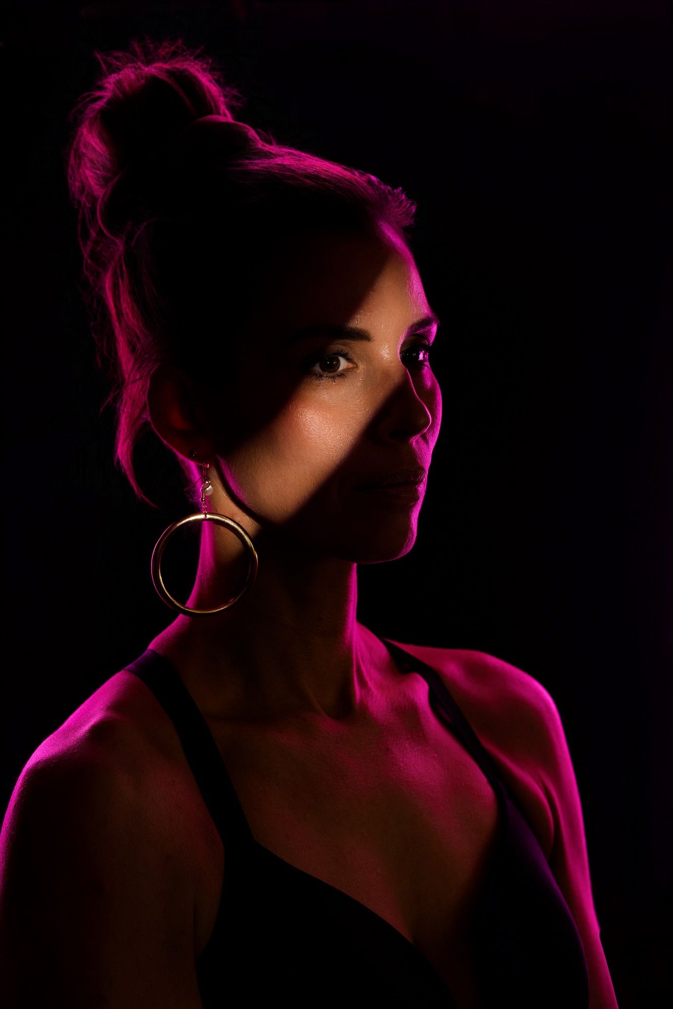 Woman with hair bun and gold hoop earring lit by dramatic pink light against dark background.