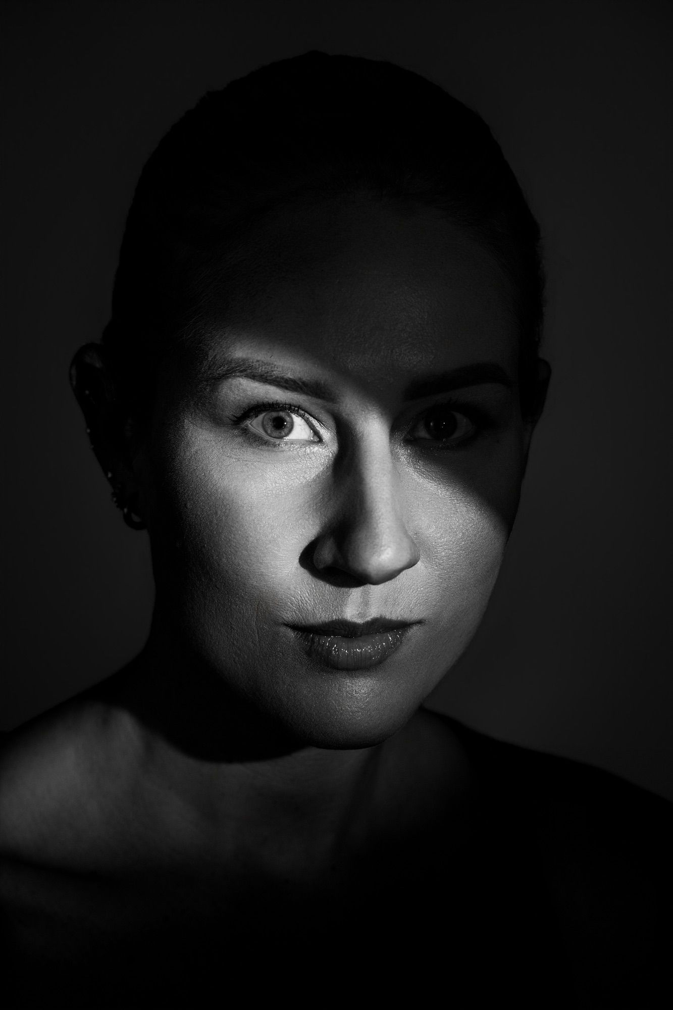 Black and white dramatic portrait of a woman with shaved head gazing intensely at camera in low-key lighting. Editorial fine art portrait