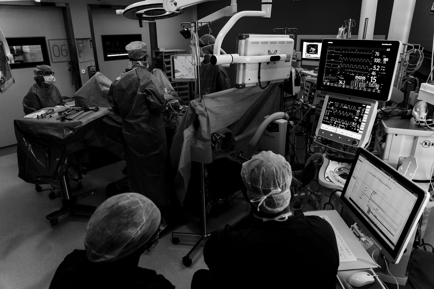 Black and white photo of surgeons operating in a modern hospital operating room with monitors displaying vital signs.
