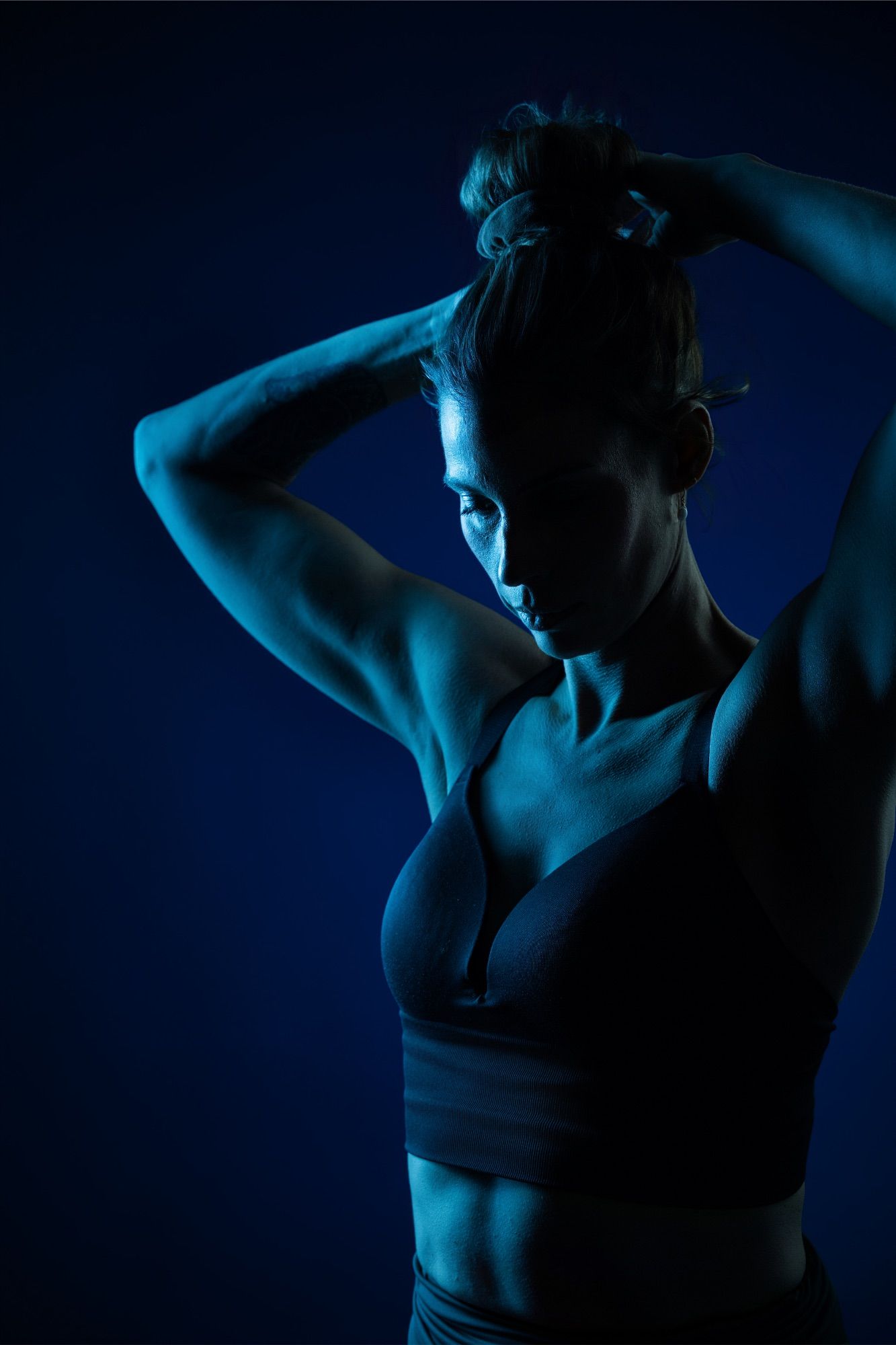 Athletic woman in a sports bra poses with hands in hair against a dark blue background in dramatic lighting.