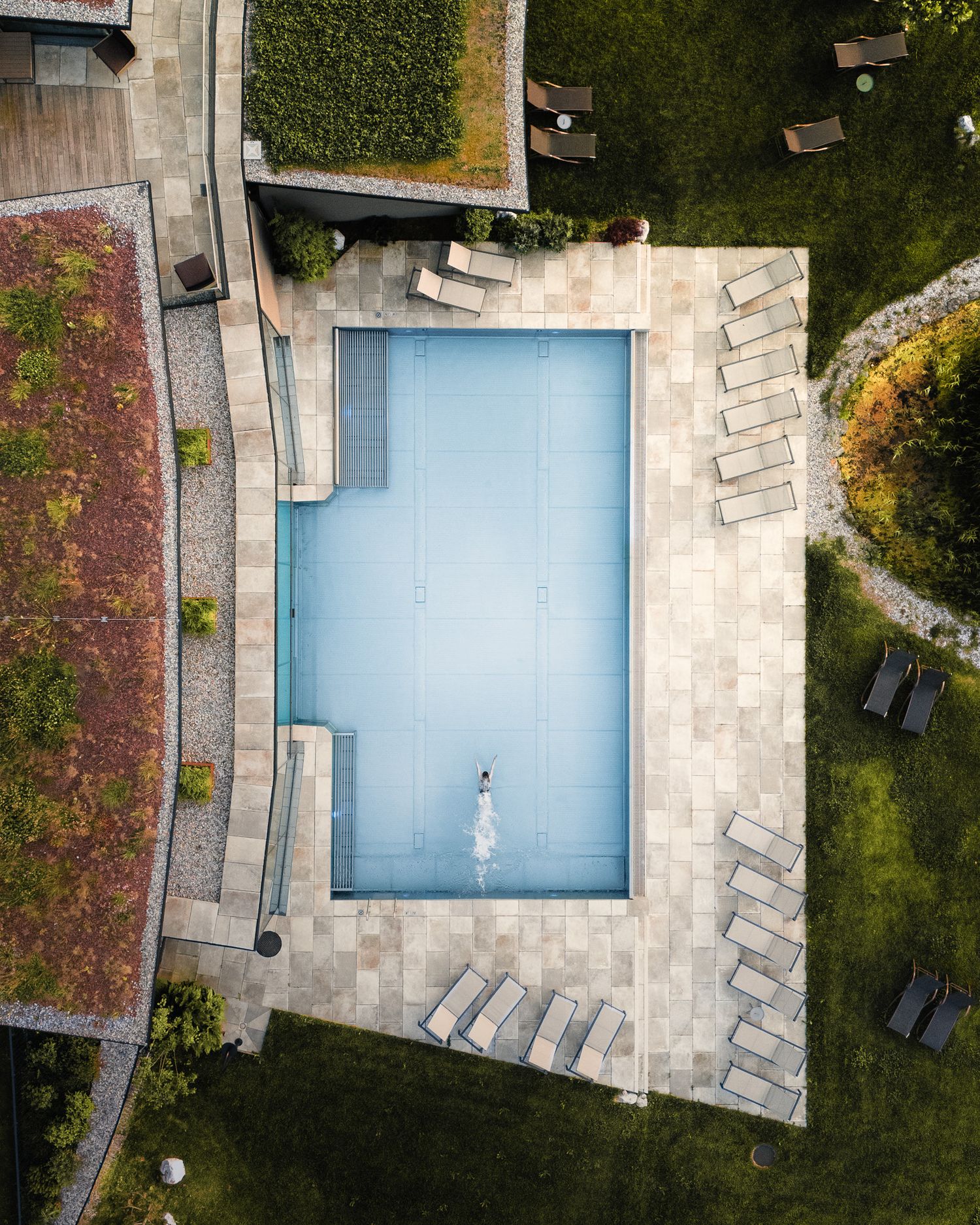 Ein Top Down Bild aus der Luft aufgenommen. Ein Pool mit einer schwimmenden Person darin. Das blaue Wasser wird von saftiger Wiese umrahmt.
