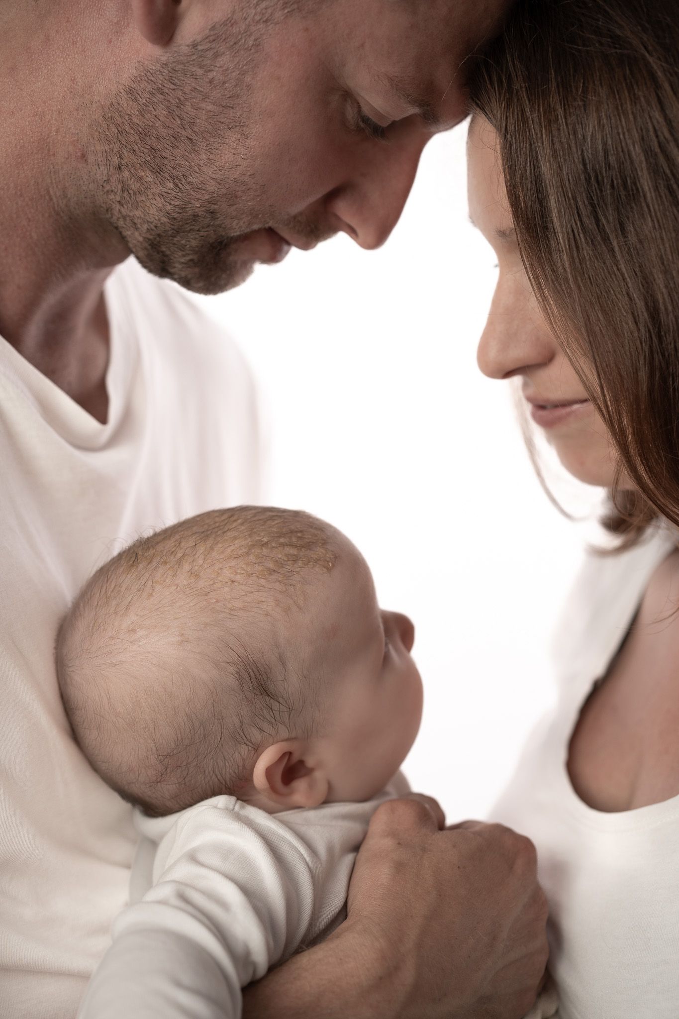 Eine Familie mit einem Baby in der Mitte auf dem Arm. Die Eltern haben die Köpfe zu einander geneigt und schauen hinab aufs Baby. High Key