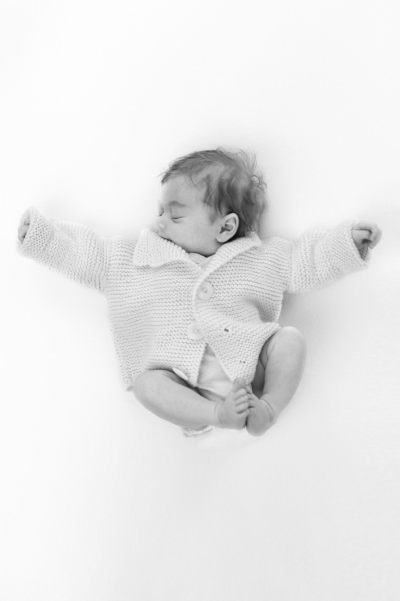 Ein Neugeborenes liegt auf dem Rücken mit angezogenen Beinen und streckt die Arme aus. Es trägt einen Strickpulli. Süßes schwarz-weiß-Foto.