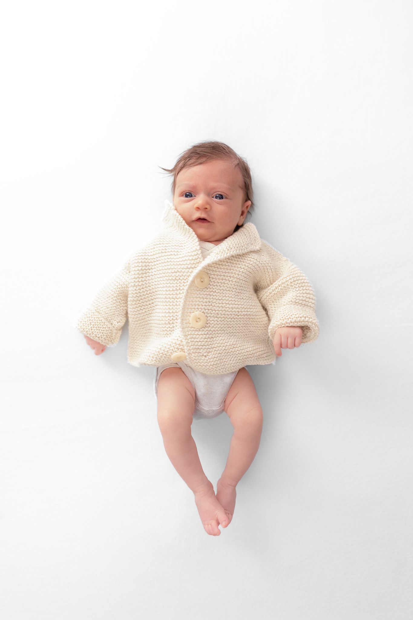 Ein Neugeborenes liegt auf dem Rücken. Es hat eine beige Strickjacke an. High Key Fotografie.