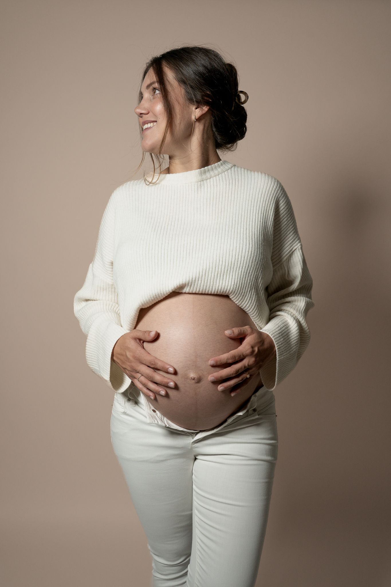 eine strahlende Schwangere in weißer Kleidung die stolz ihren Babybauch umfasst und lächelnd zur Seite schaut.