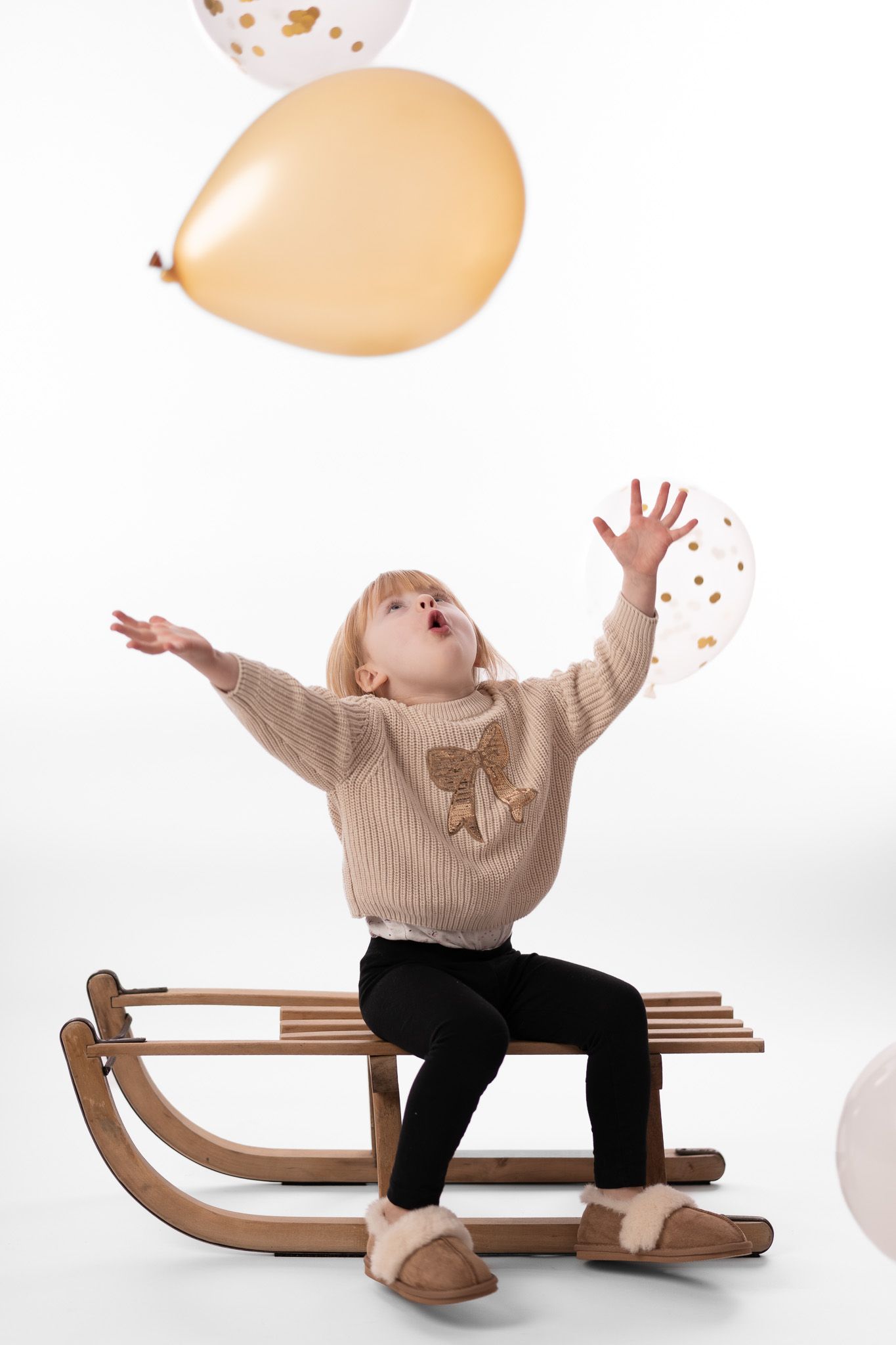 Ein Kleinkind sitzt auf einem Schlitten und spielt mit Luftballons. Es streckt sich in die Höhe um einen Ballon zu fangen. High Key