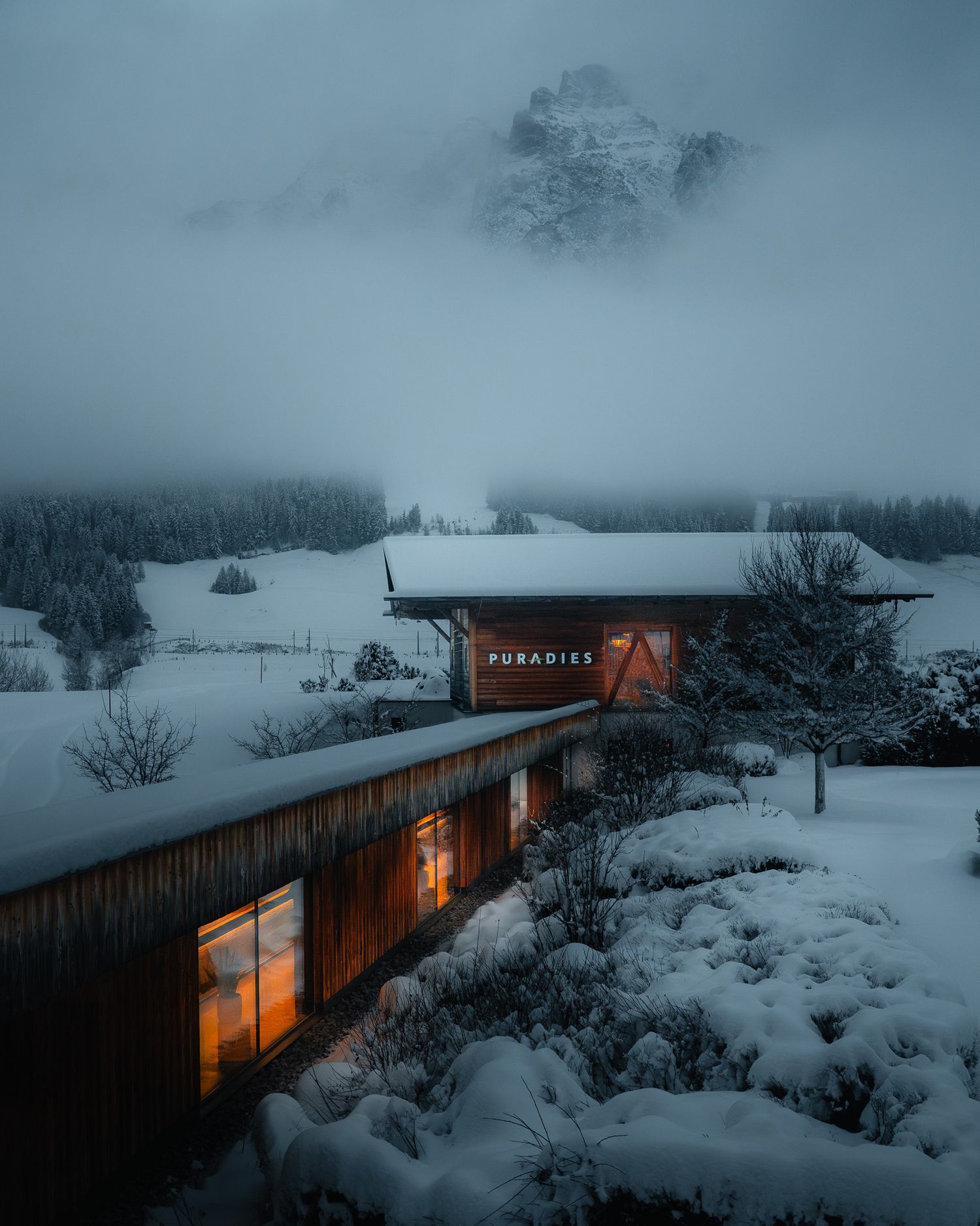 Eine Schneelandschaft im Morgengrauen. Der Schnee bedeckt ein Saunahaus, indem ein warmes Licht brennt. Im Hintergrund Bergpanorama im Nebel