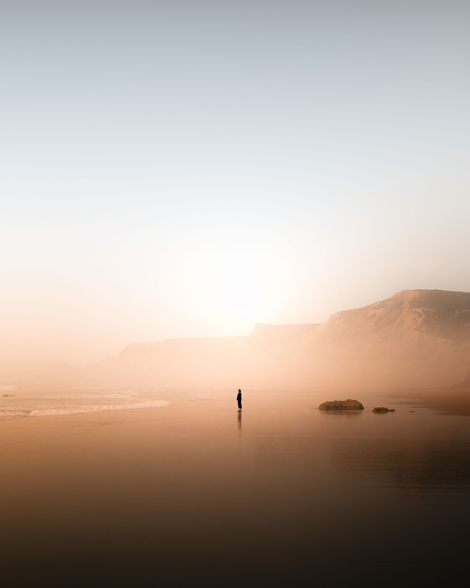 Ein beeindruckender Sonnenuntergang der den Dunst am Strand von Portugal in warme Töne färbt und eine perfekt positionierte Person im Bild.