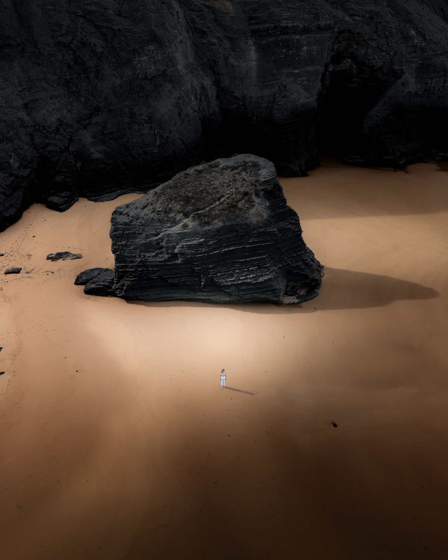 Ein minimalistisches Landschaftsbild aus Portugal, auf dem eine Frau an einem schwarzen Felsen am goldenen Strand vorbeigeht.
