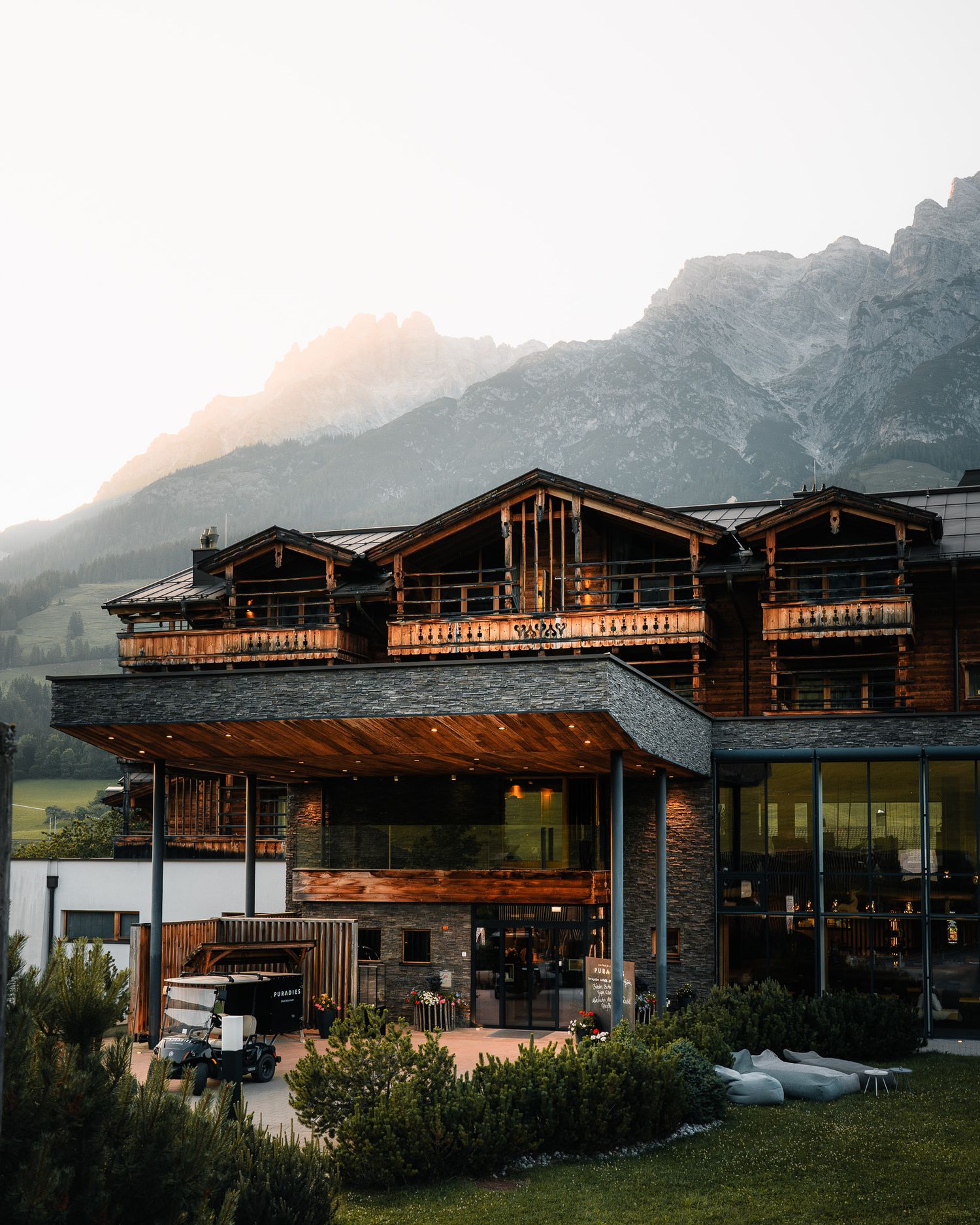 Ein einladendes Hotel mit moderner Holzfassade und großen Glaselementen, im Hintergrund die Berge, im Vordergrund saftige grüne Wiesen.