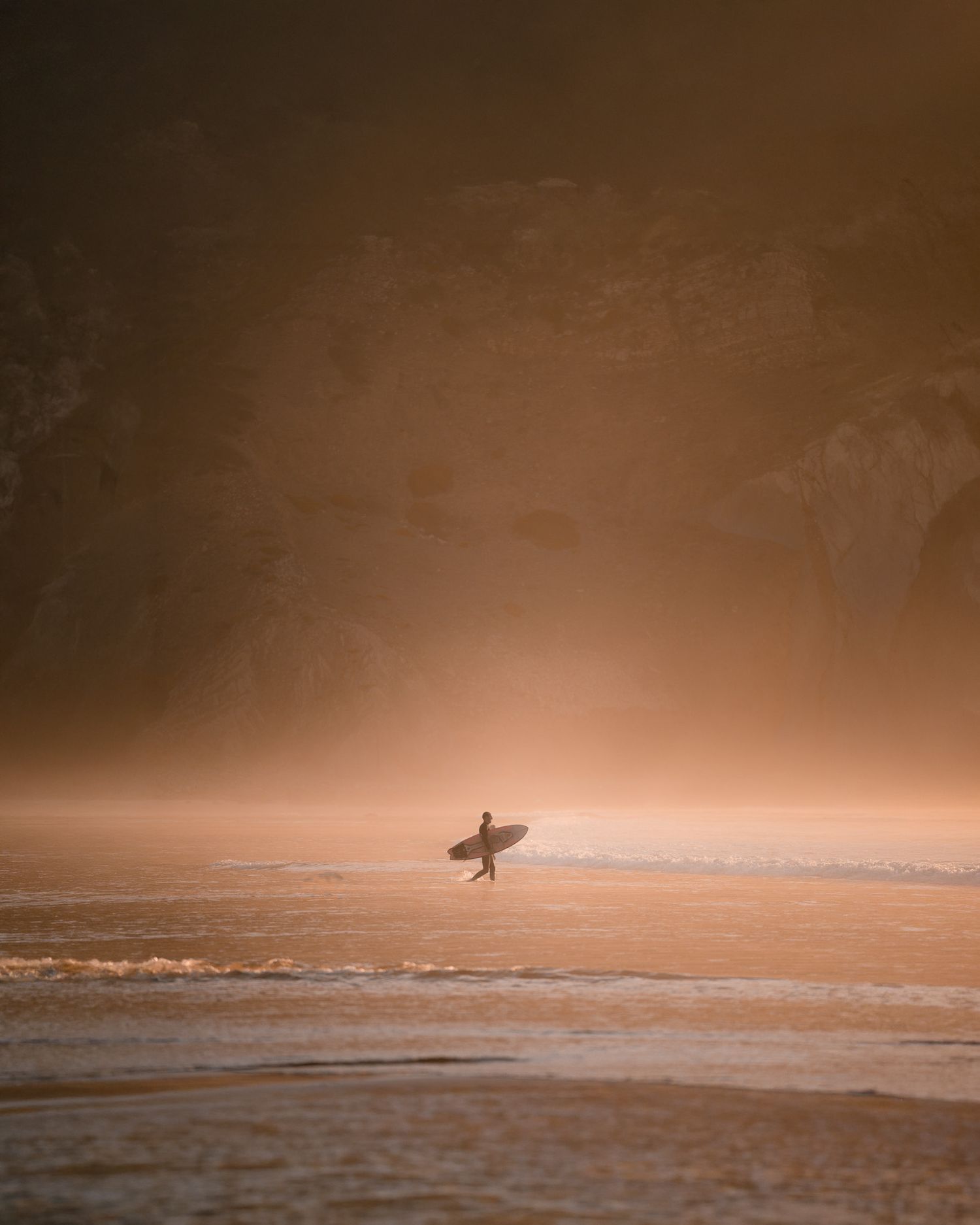 Ein Surfer auf dem Weg zurück ins Meer mit seinem Surfbrett unter dem Arm. Er wird von der Abendsonne in warmes, goldenes Licht getaucht.