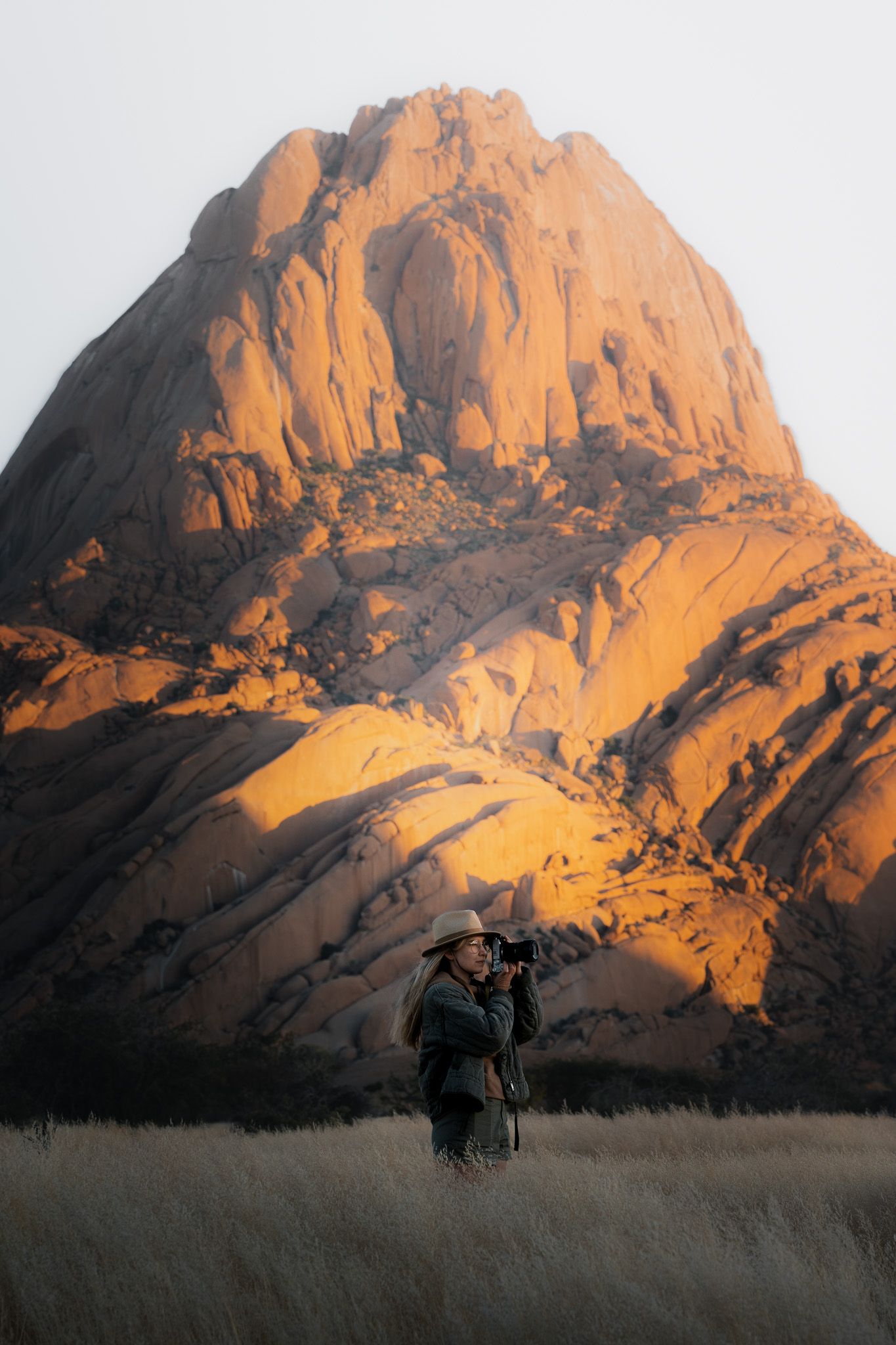 Eine Frau steht in hohem Gras und fotografiert etwas in der Ferne. Im Hintergrund ein golden angestrahlter Fels in Spitzkoppe, Namibia.