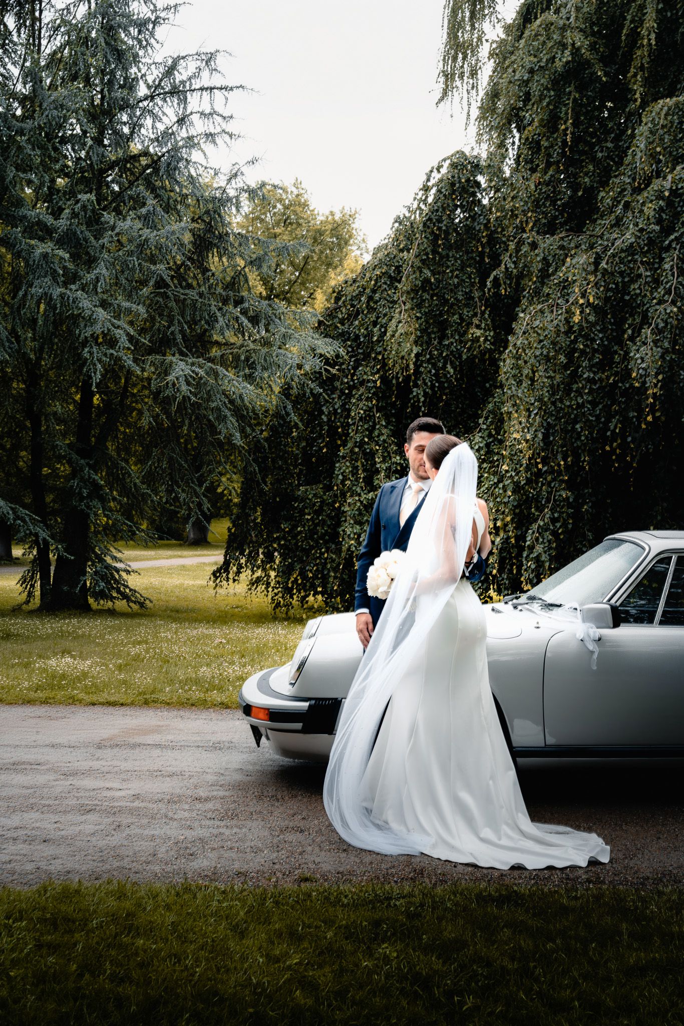 Ein Brautpaar steht im Park vor einem grauen Porsche Oldtimer und sieht sich intensiv an.
