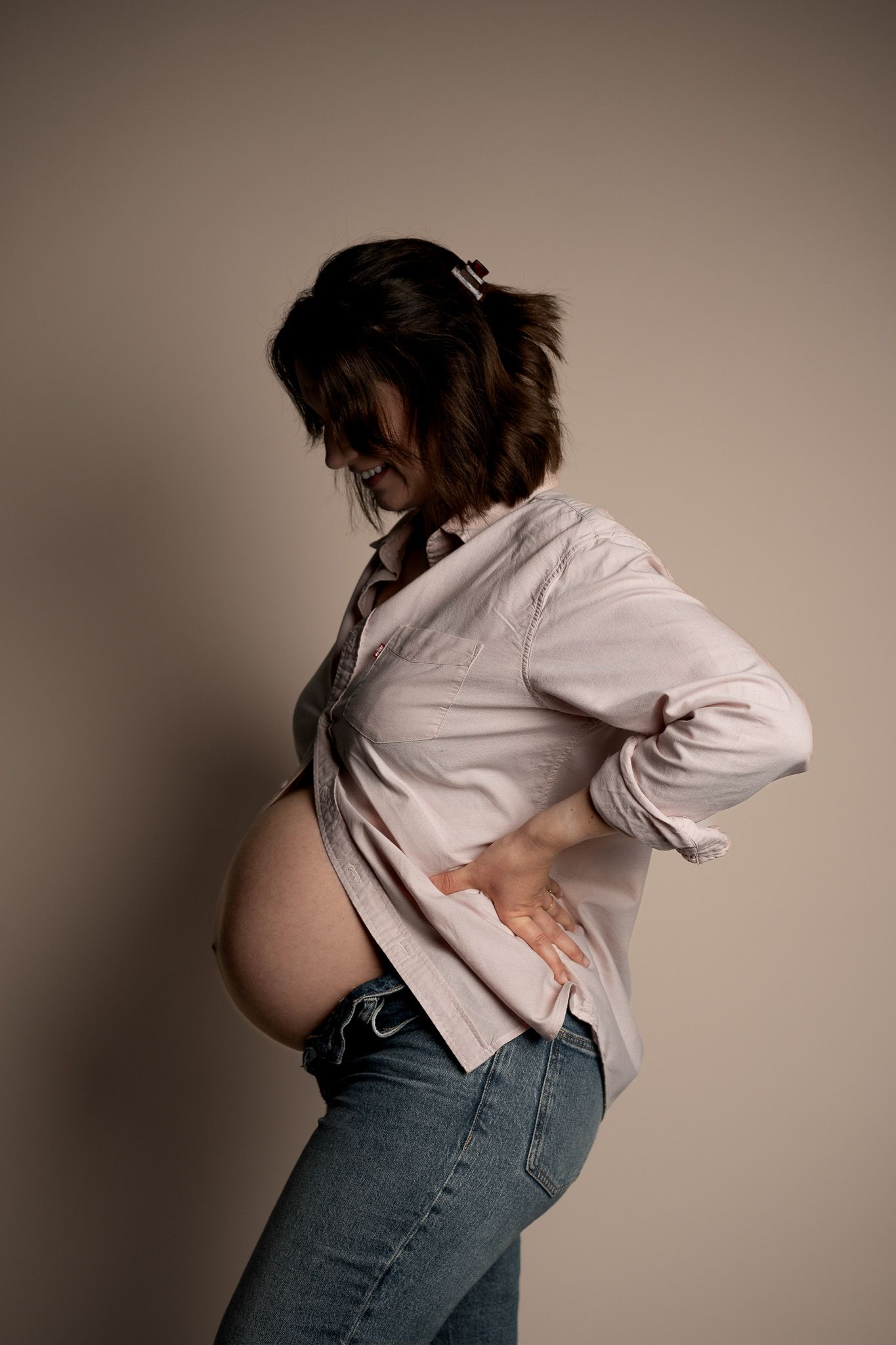 Eine Schwangere Frau im Profil mit einem Hemd an aus dem ihr Babybauch hervorschaut. Sie steht im Profil da und schaut auf ihren Bauch.
