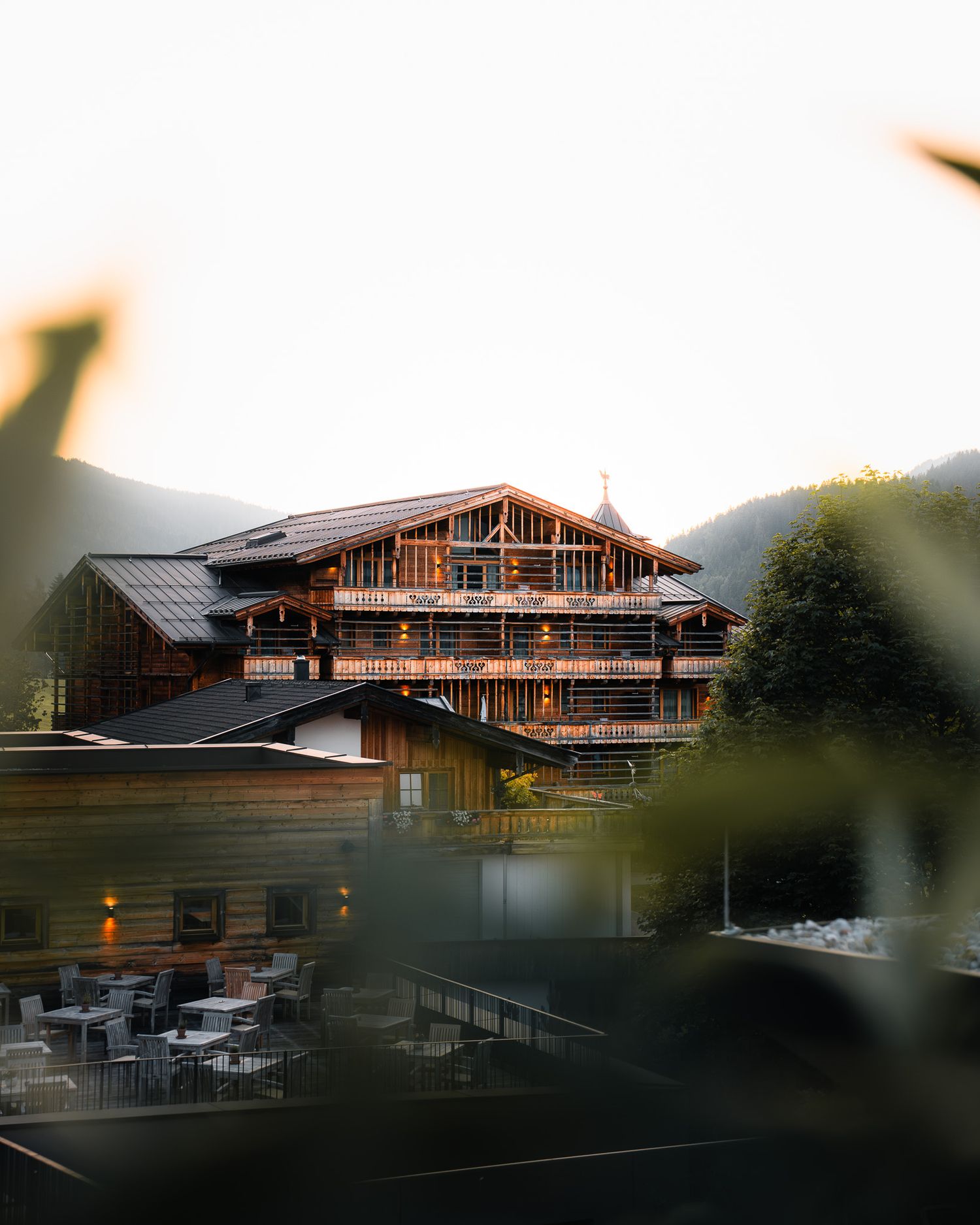 Ein modernes Wellness-Hotel mit Holzfassade, aus der Ferne fotografiert, eingerahmt von grünen Blättern eines Strauches im Vordergrund.